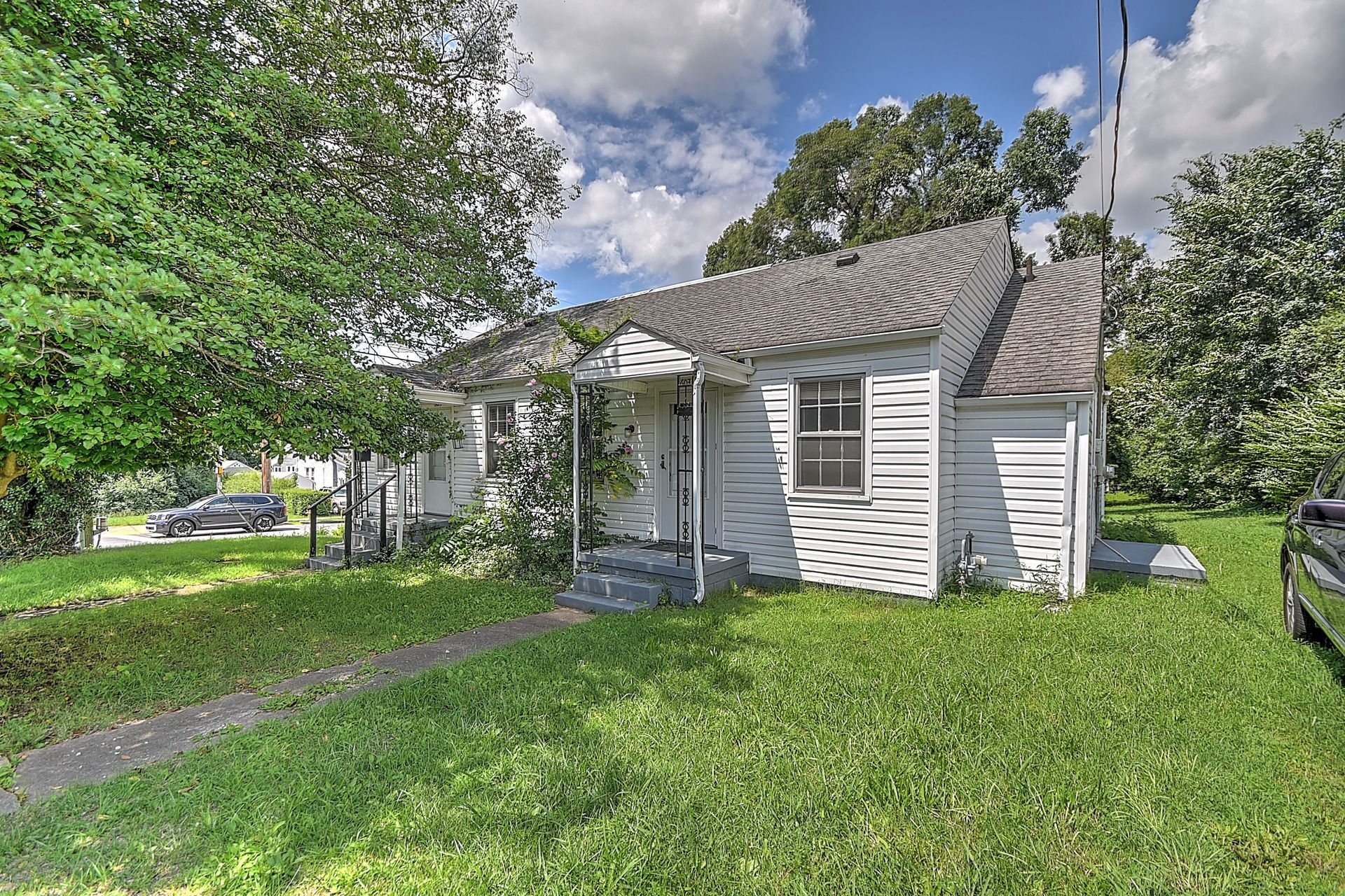Small white house with a front porch, gray roof, and green yard under a partly cloudy sky