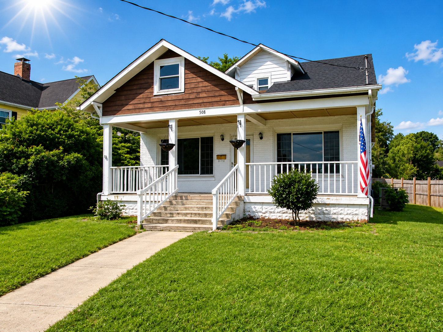 Small beige house with a front porch, green lawn, and American flag on a sunny day