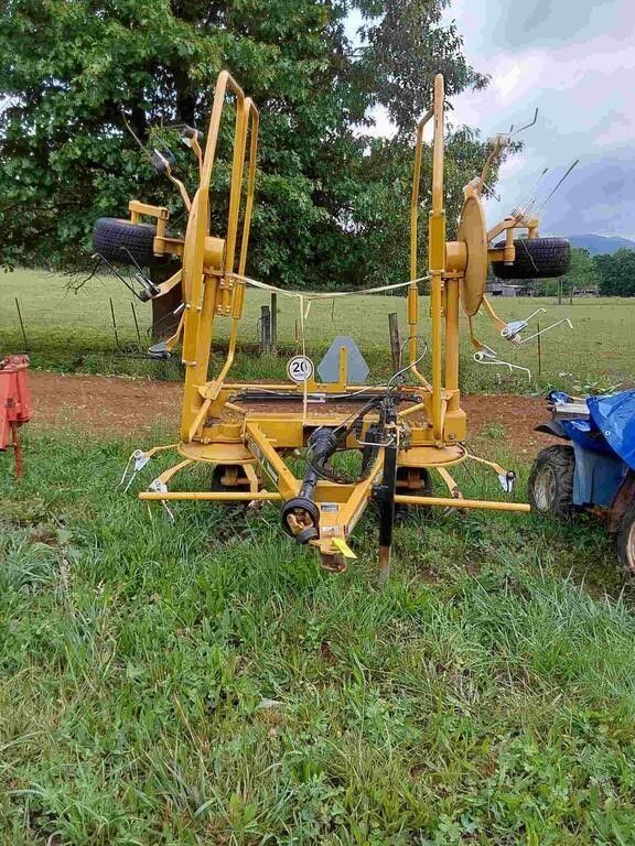 Yellow agricultural machine in a grassy field, with trees and a blue tractor nearby
