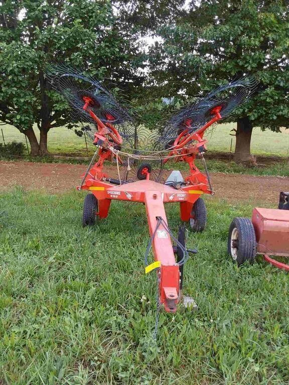 Red agricultural machine with orange and black parts on grass near trees, with a cart wheel visible at right.