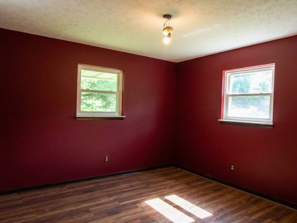 Empty room with dark red walls, wood floor, and two windows letting in sunlight.