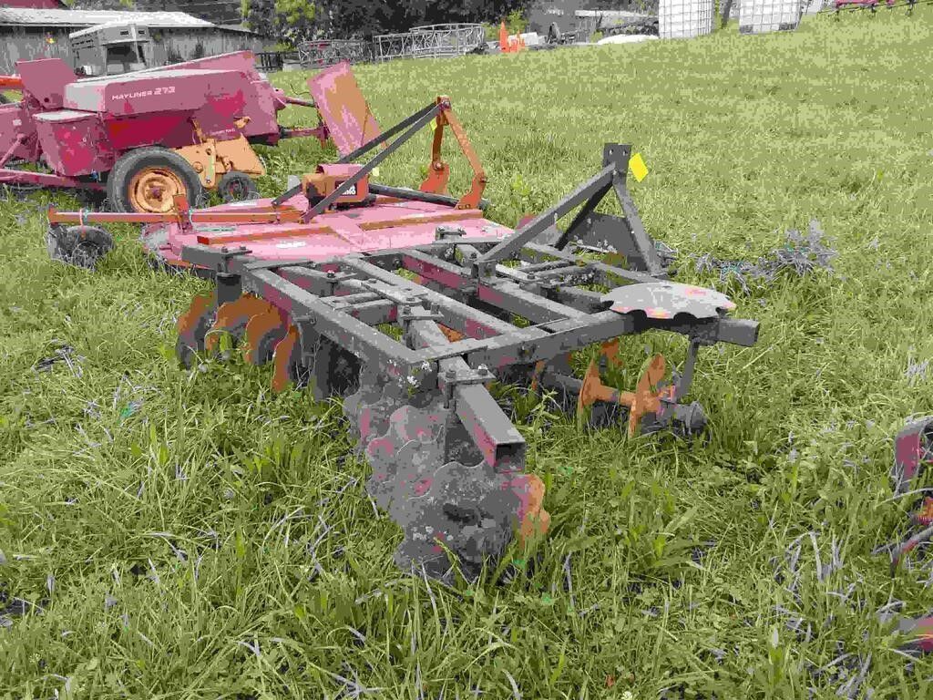 Red agricultural tillage implement in a grassy field, with a tractor visible behind it