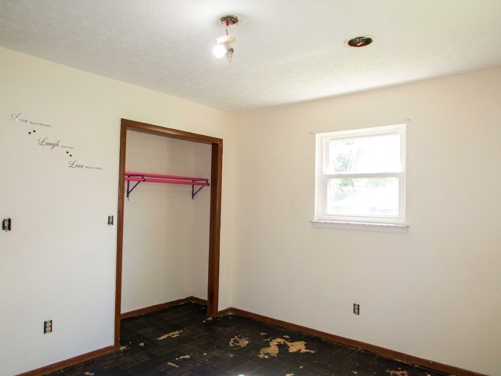 Empty bedroom with beige walls, closet opening, window, and dark carpet remnants on the floor
