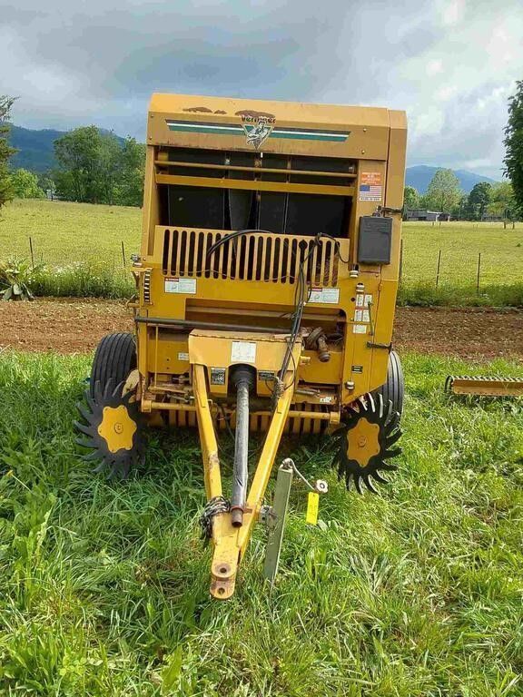 Yellow agricultural machine parked in a grassy field with farm machinery and hills in the background