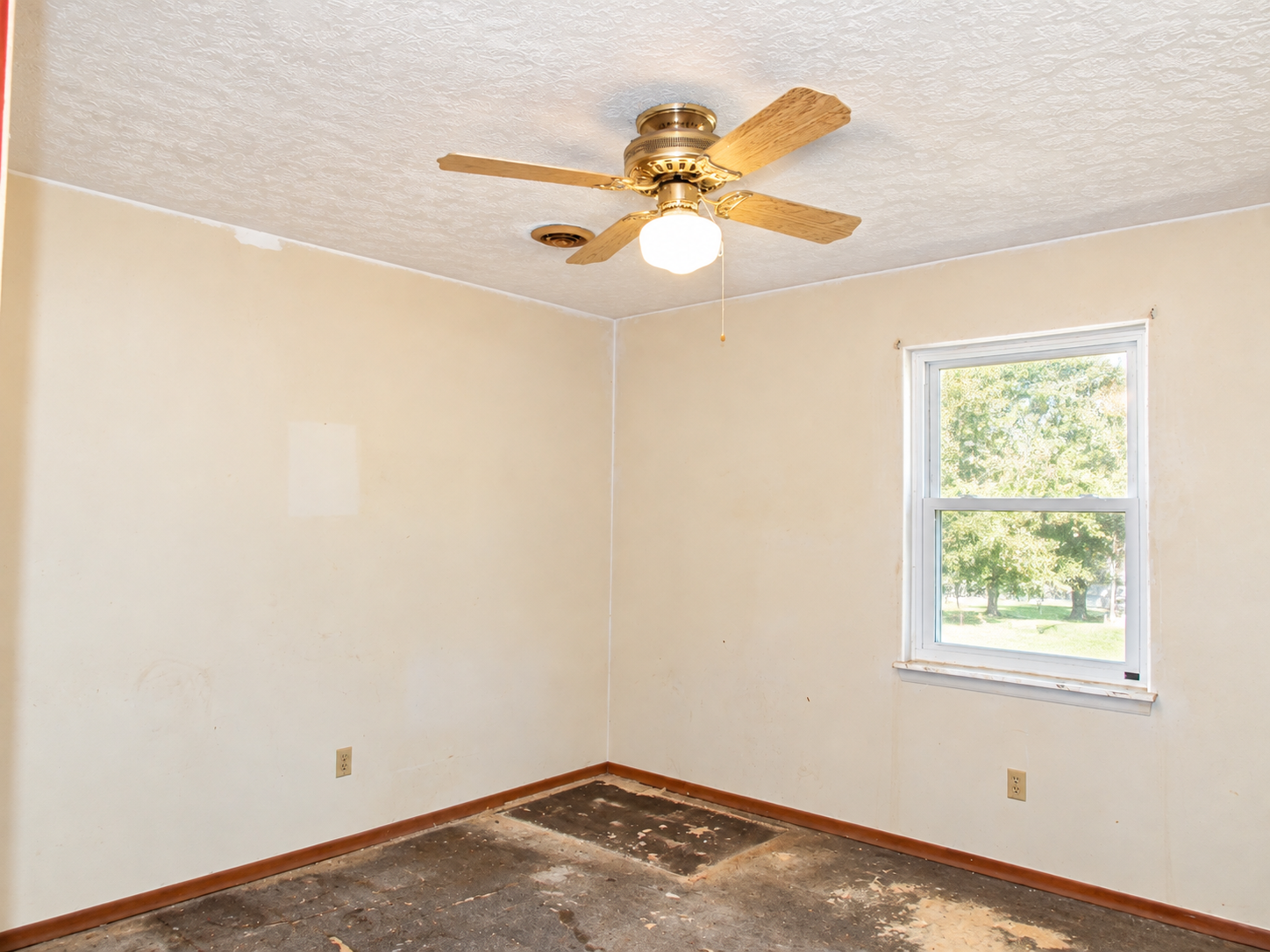 Empty beige room with ceiling fan, white window, and bare floor patch near one corner
