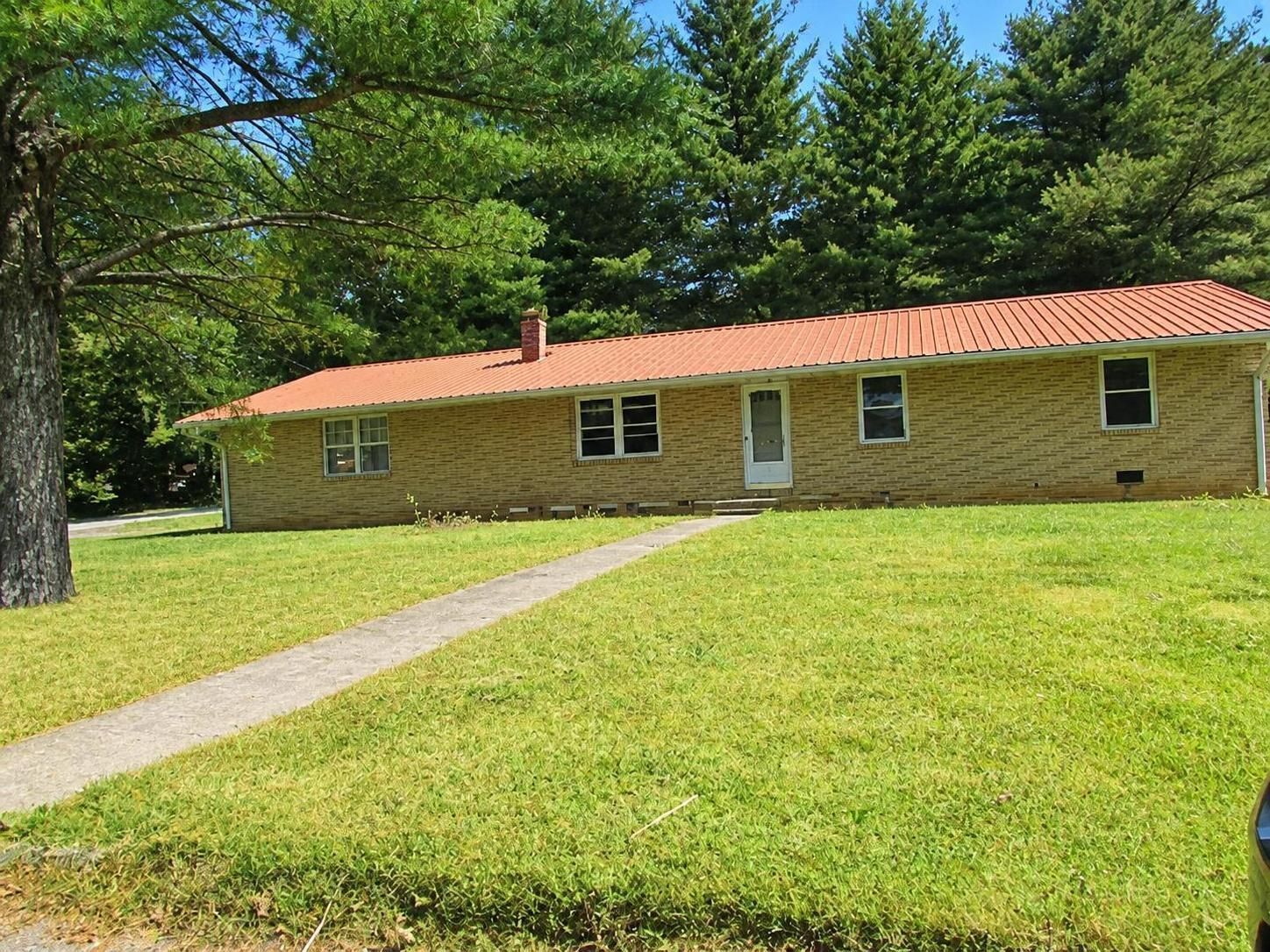 Single-story tan brick house with red roof, front lawn, and curved driveway surrounded by trees