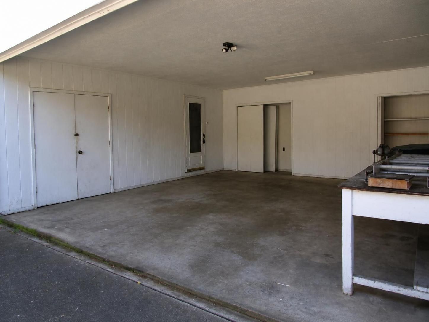 Empty carport with concrete floor, white walls, and a small workbench on the right.