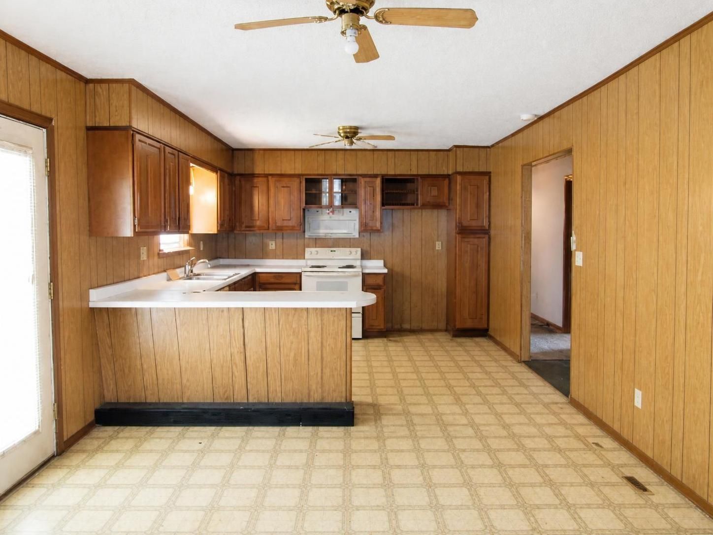 Empty vintage kitchen with wood paneling, white counters, and beige patterned linoleum floor.
