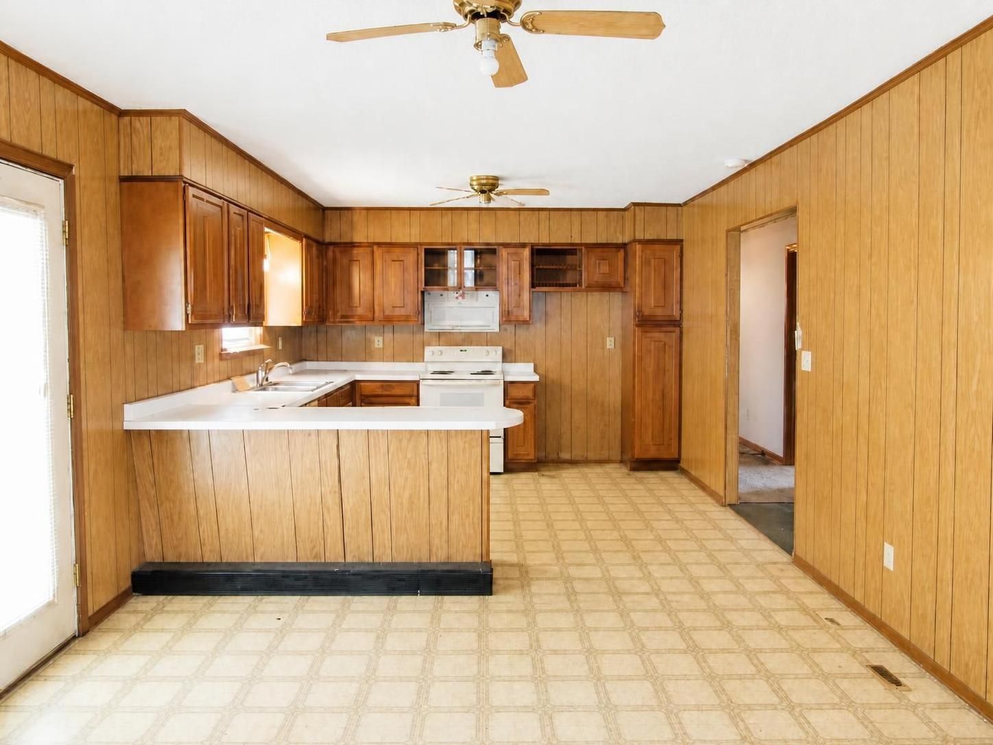 Empty wood-paneled kitchen with white counters, tan tile floor, and ceiling fans