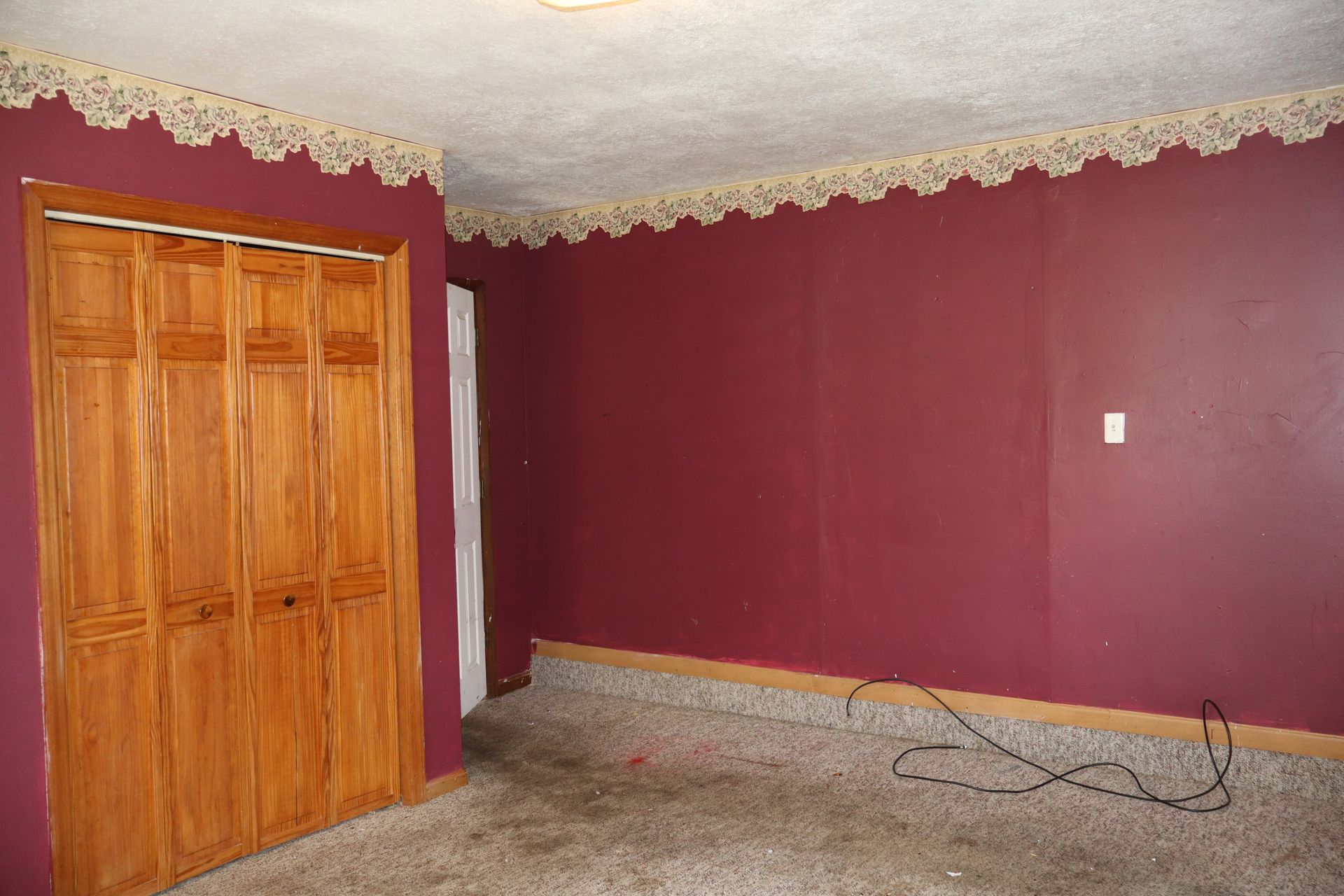 Empty room with burgundy walls, wooden closet doors, patterned ceiling trim, and a carpeted floor with cables.