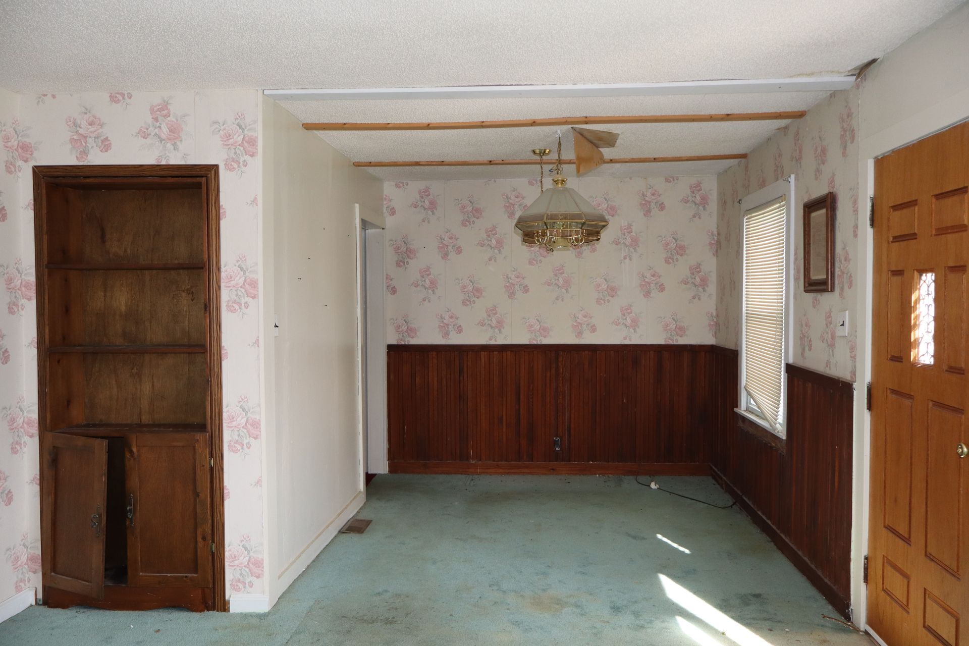 Empty room with floral wallpaper, wood paneling, teal carpet, and a wooden door on the left and right.
