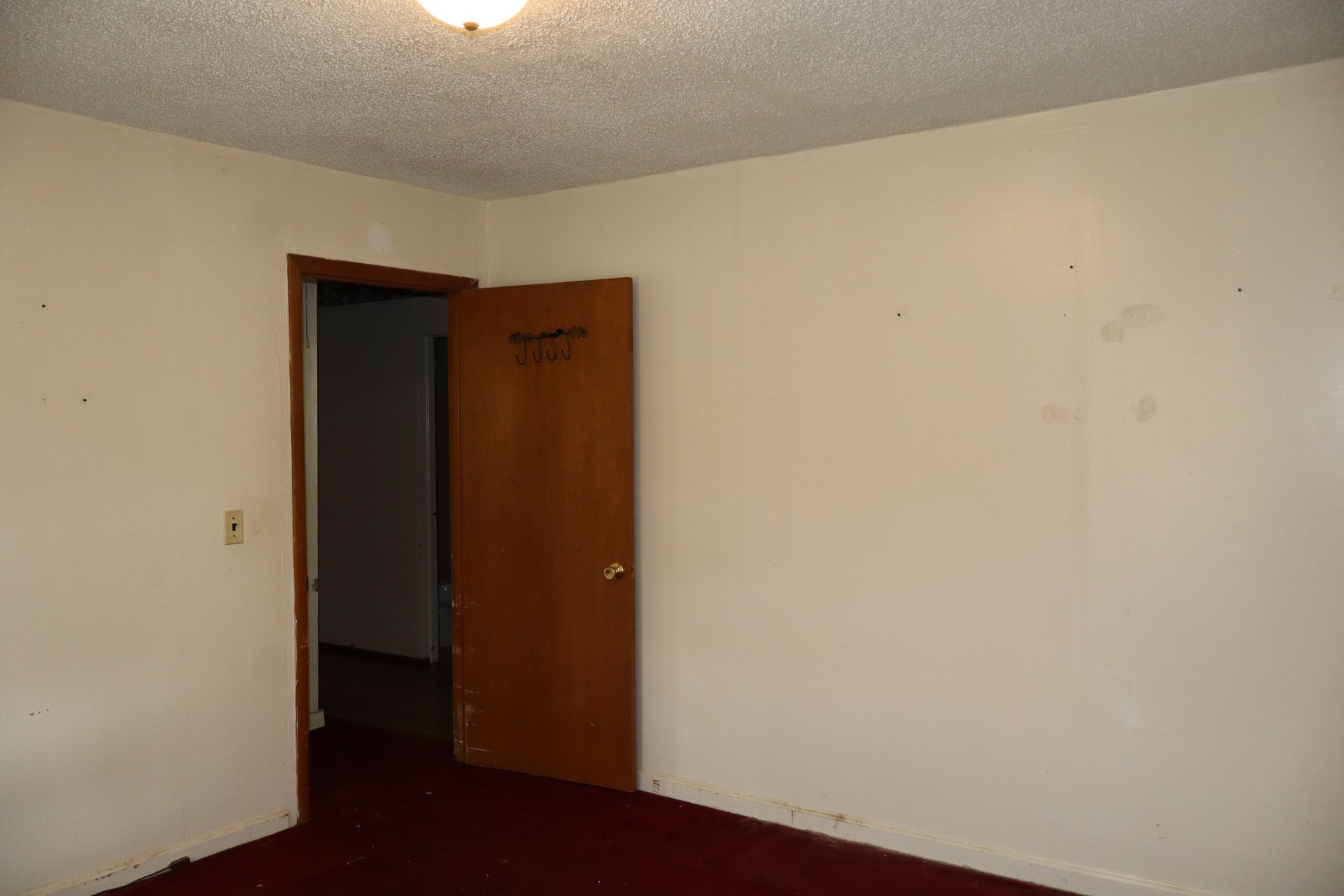 Empty room with white walls, maroon carpet, and an open wooden door leading to a dark hallway