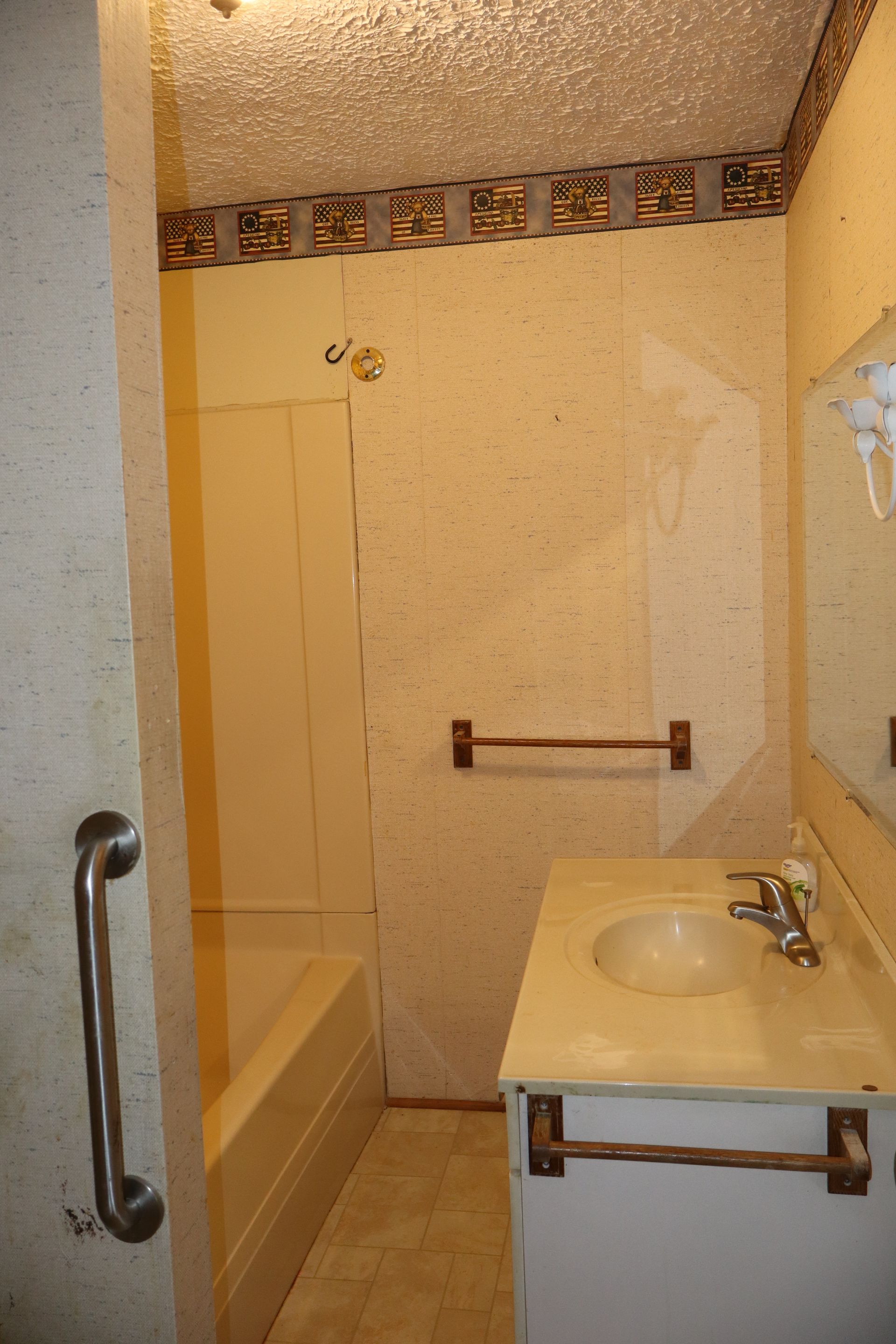 Small beige bathroom with a bathtub, sink, mirror, and grab bars.