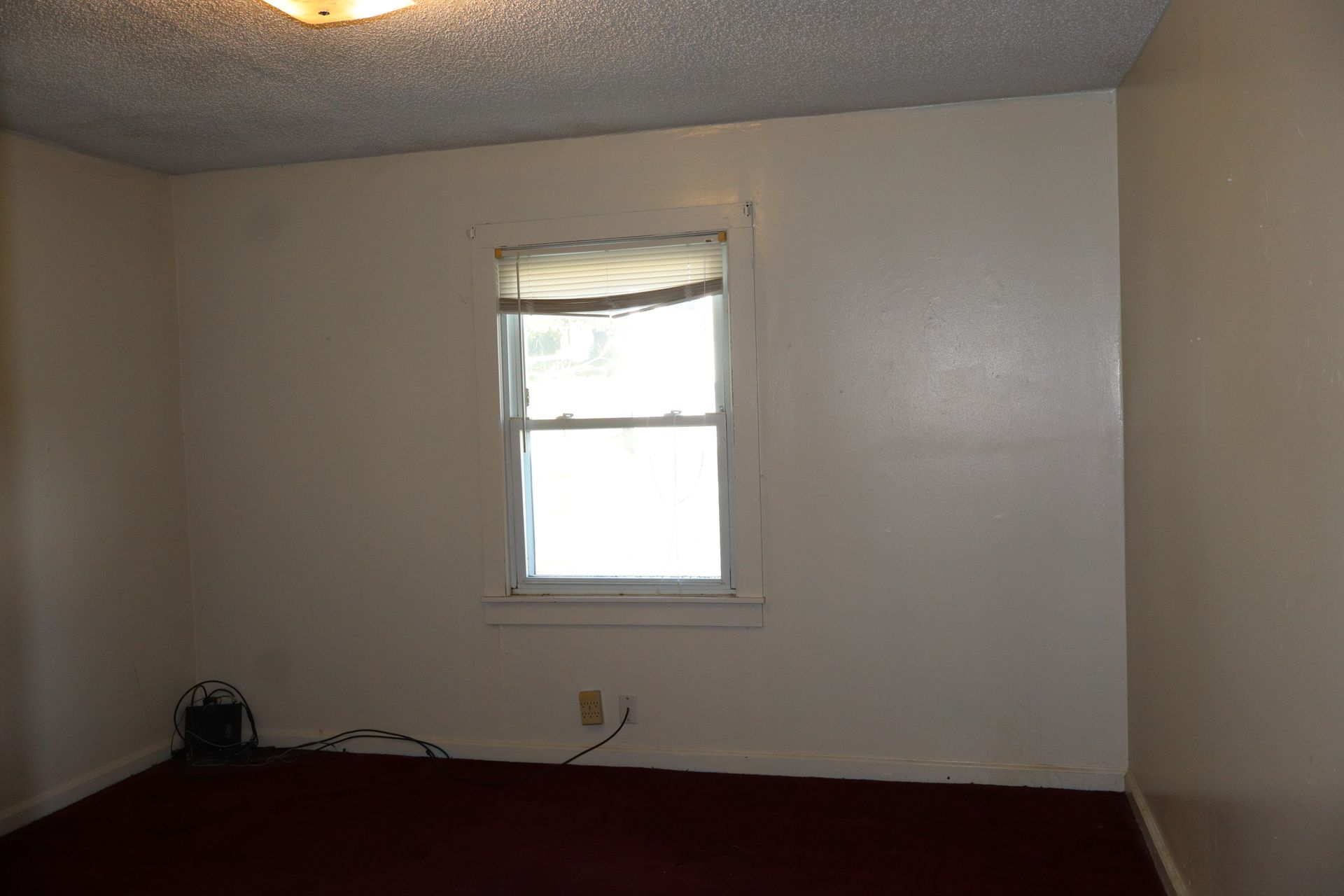 Empty room with white walls, maroon carpet, and a small window with blinds.