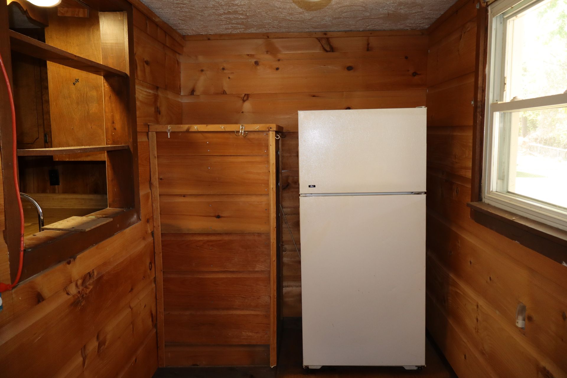 Wood-paneled kitchen with a white refrigerator, window, shelves, and wooden cabinet.