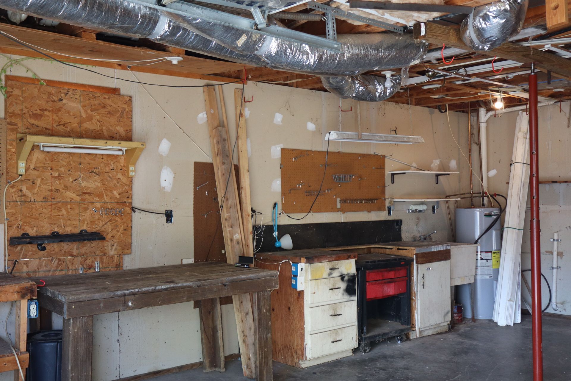 Cluttered garage interior with exposed insulation, workbench, cabinets, and a red post