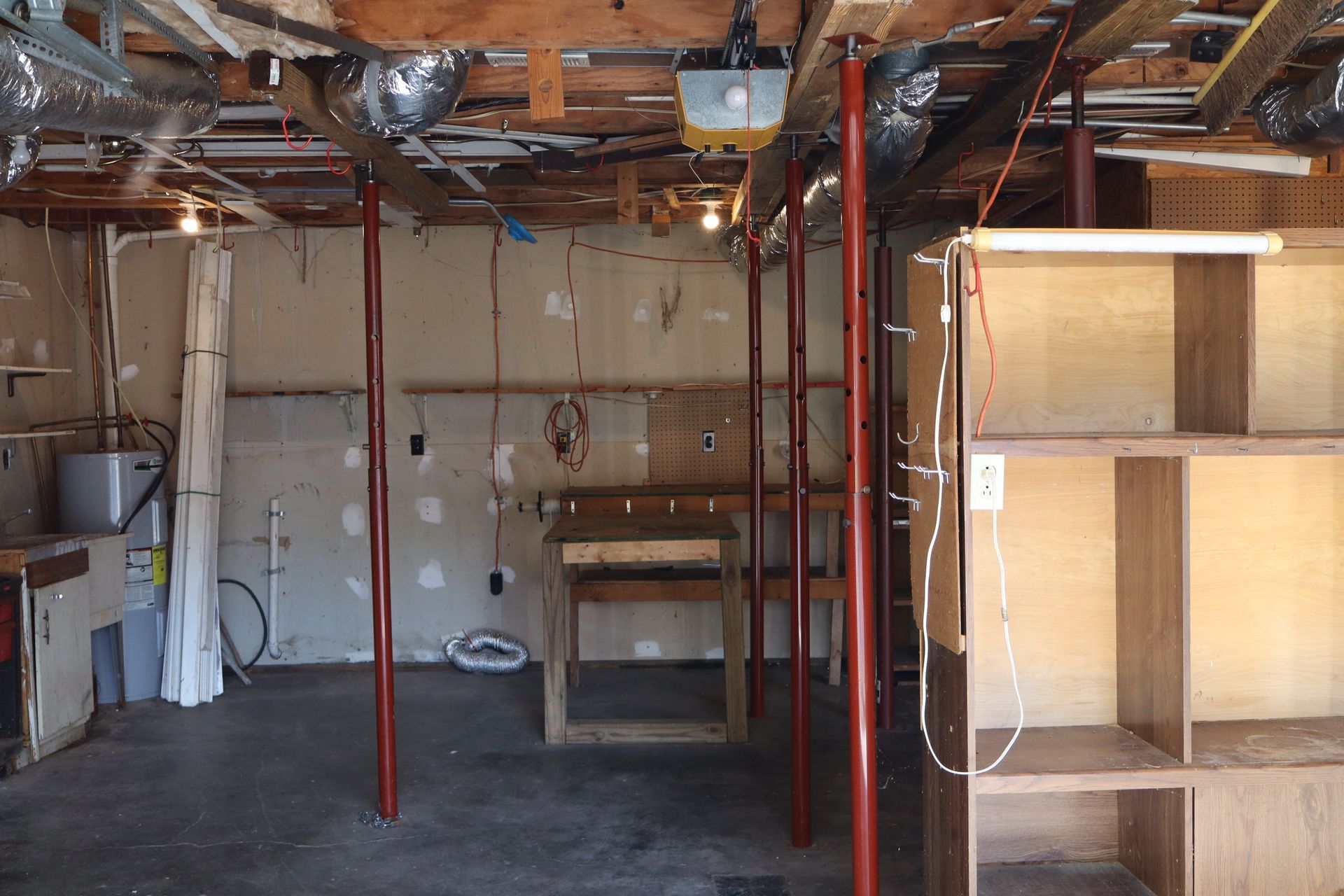 Unfinished basement with exposed beams, red support poles, utility appliances, and wooden shelving.
