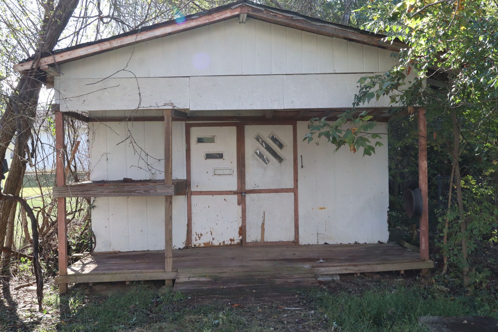 Small weathered white shed with a porch, wooden posts, and overgrown vines in a yard.