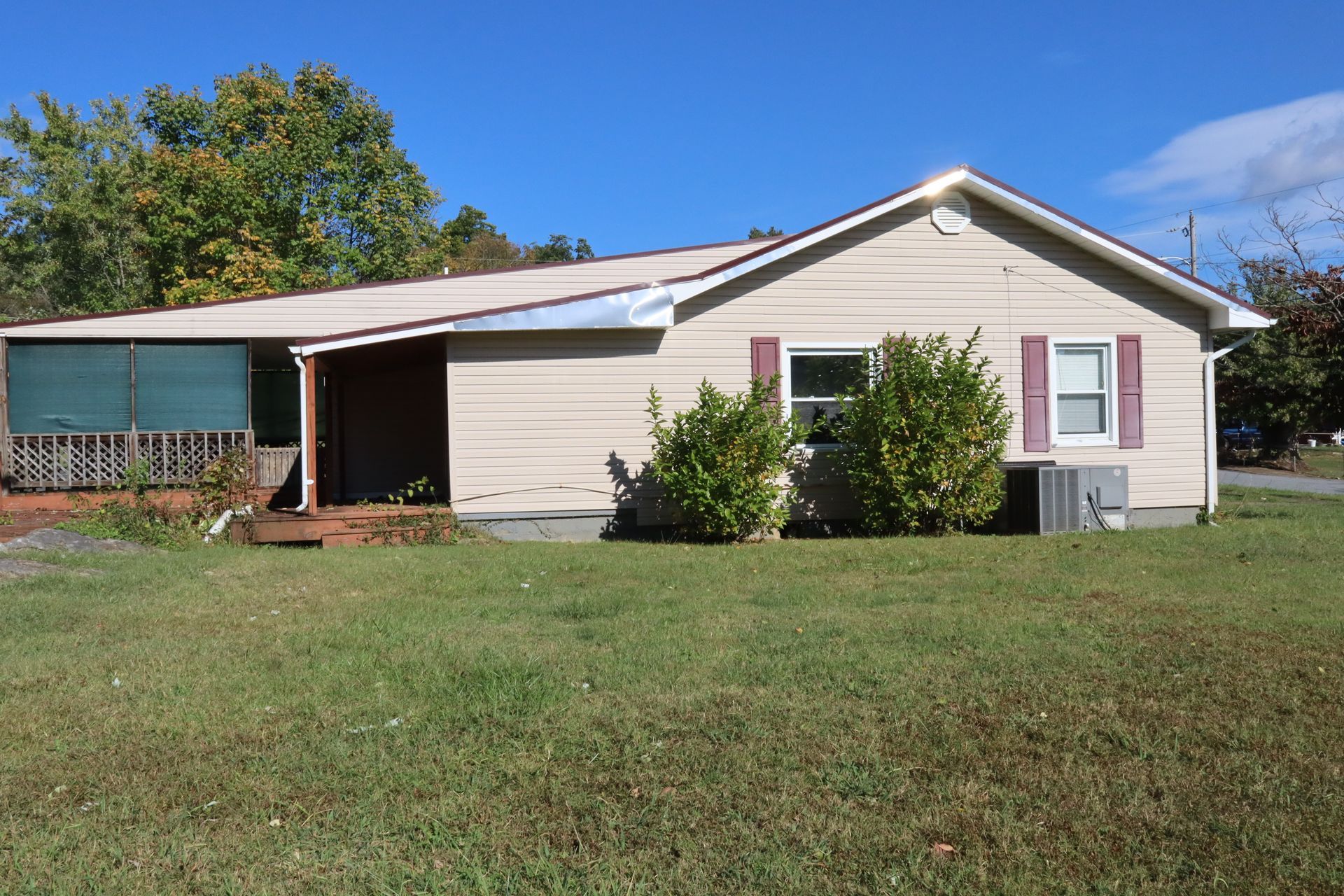 Single-story beige house with a small porch, shrubs, and a large grassy yard under a blue sky