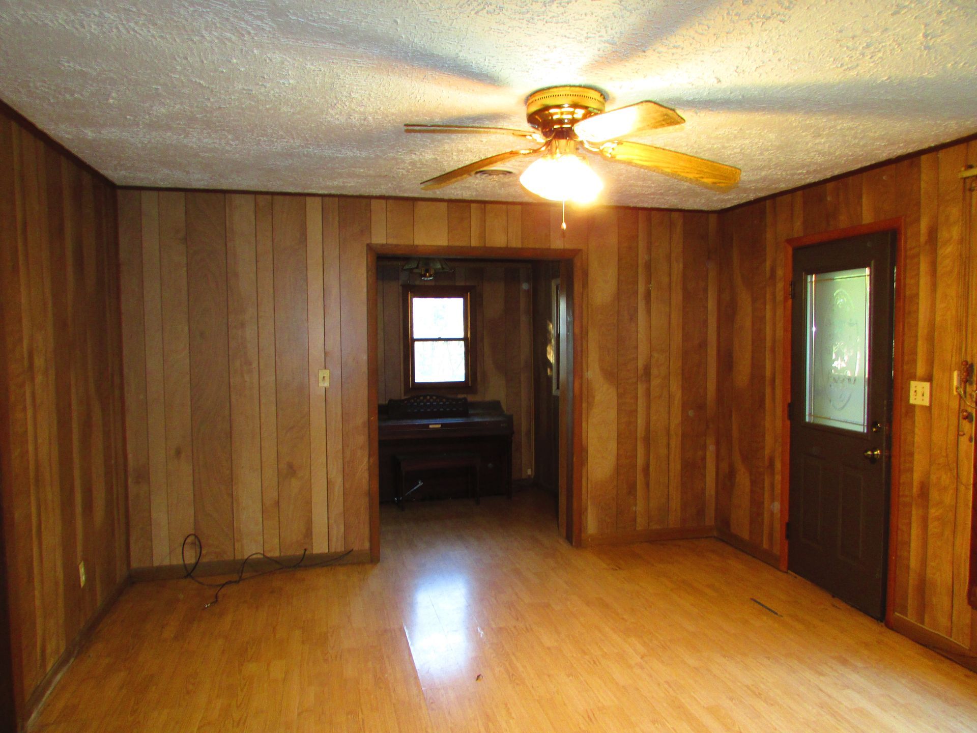 Room with wood paneling walls, wood floor, and doorway to another room with piano.