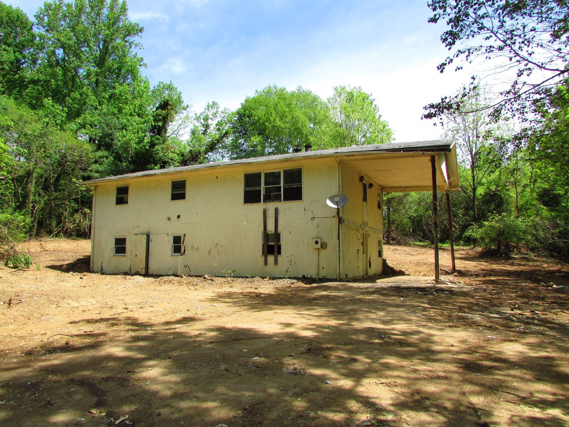 Small yellow house with a covered porch, surrounded by trees on a dirt clearing.