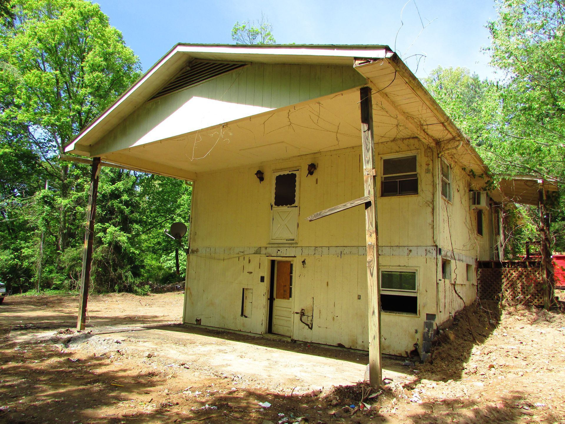 Two-story yellow house with a large overhanging roof, surrounded by trees on a dirt lot.