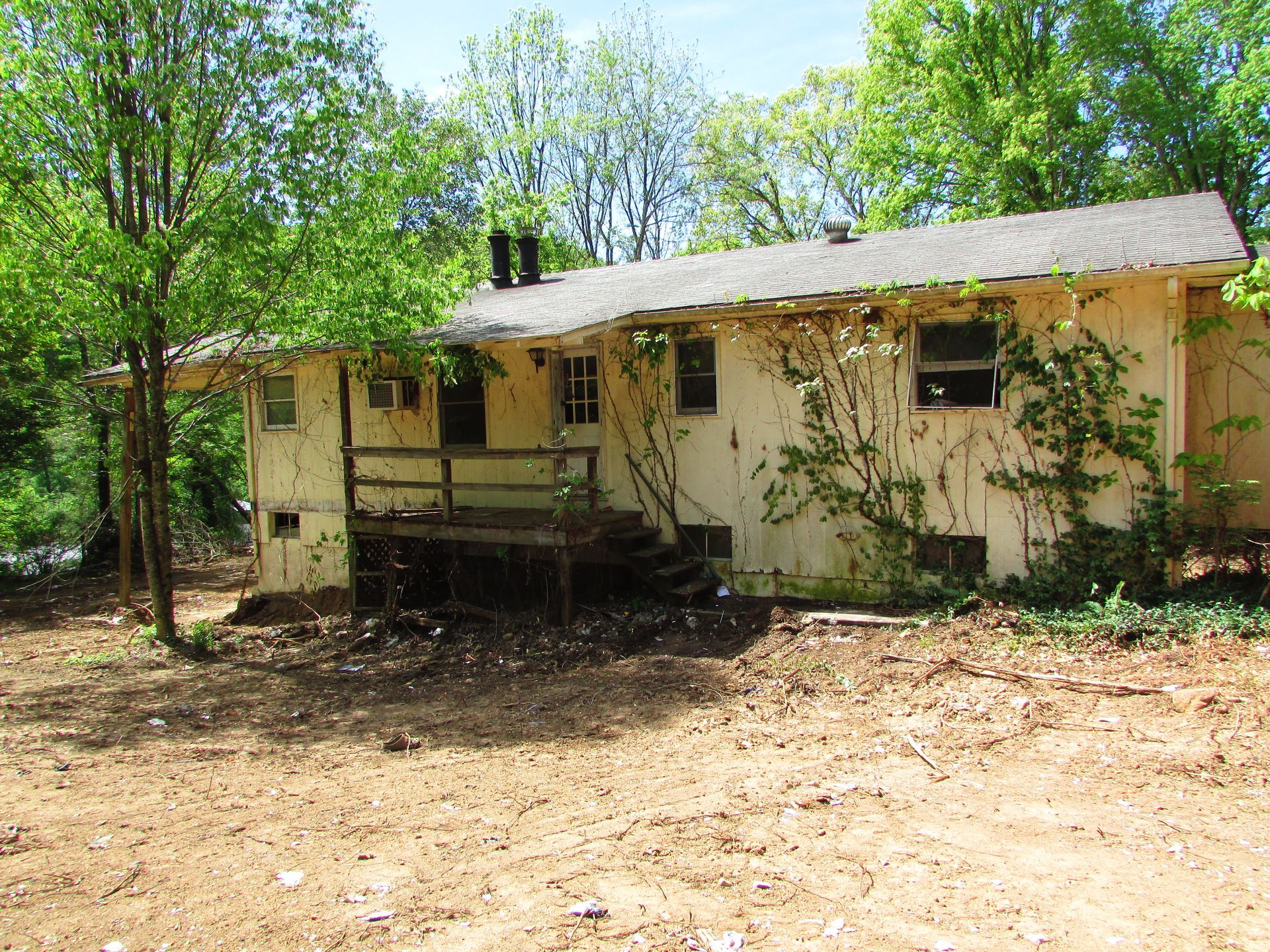 Small beige house with a front porch in a wooded yard, surrounded by trees and dry ground.