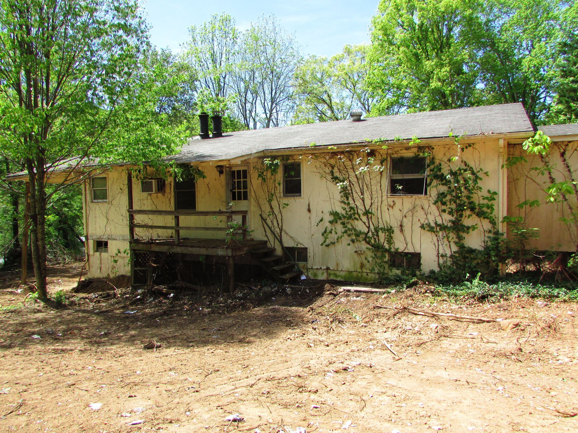 Overgrown rural house with peeling beige siding, porch, and trees in bright daylight