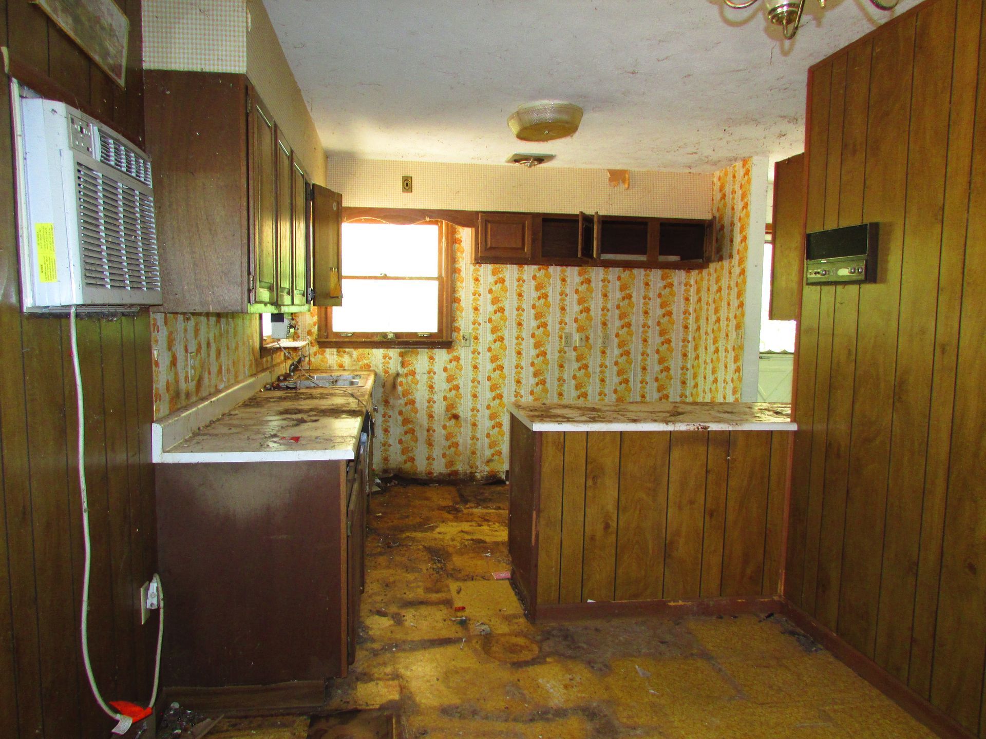 Empty old kitchen with wood-paneled walls, worn floor, and floral wallpaper around a small window.