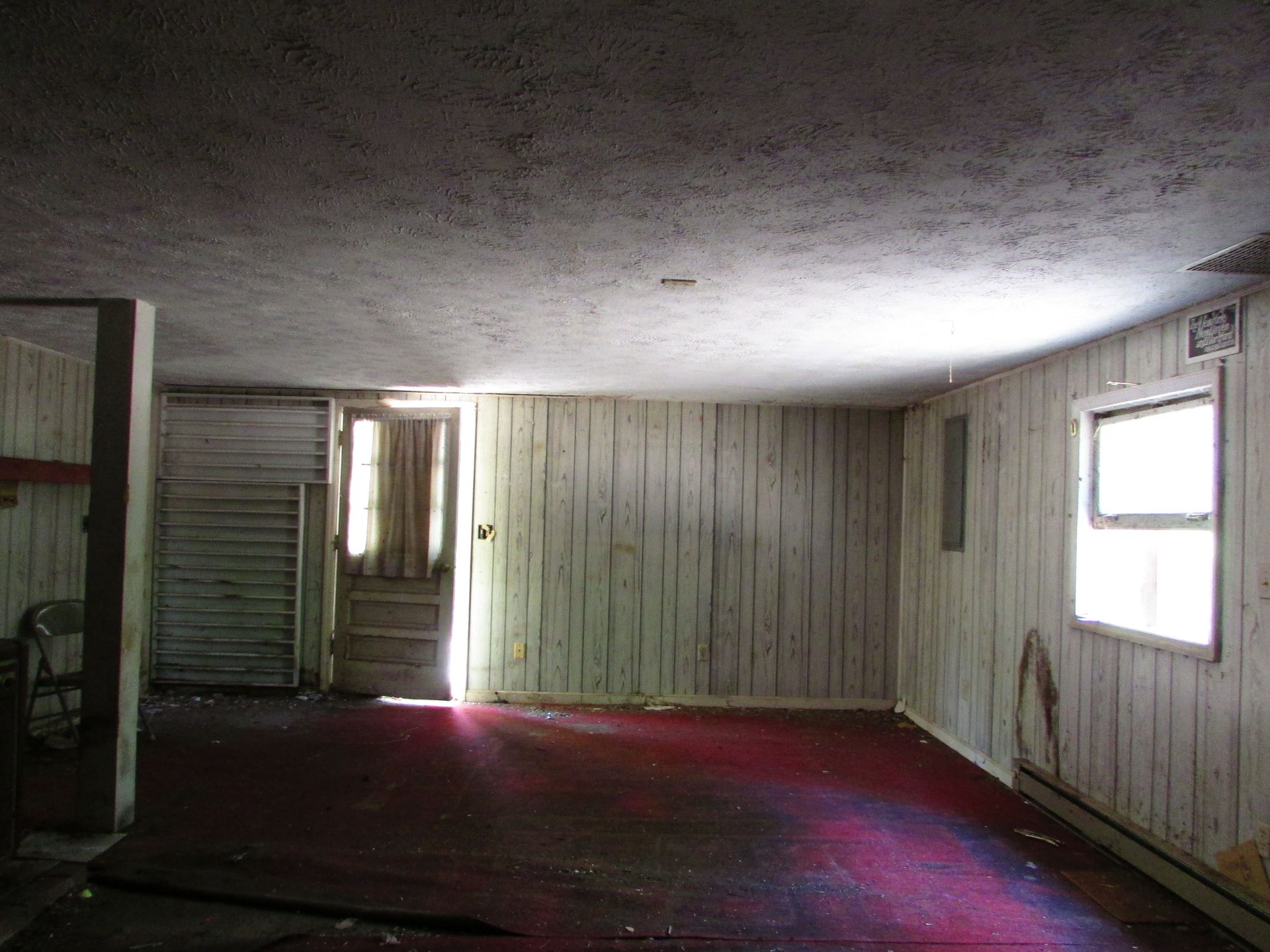 Empty worn interior room with white paneled walls, low ceiling, and red carpet near windows and doorway