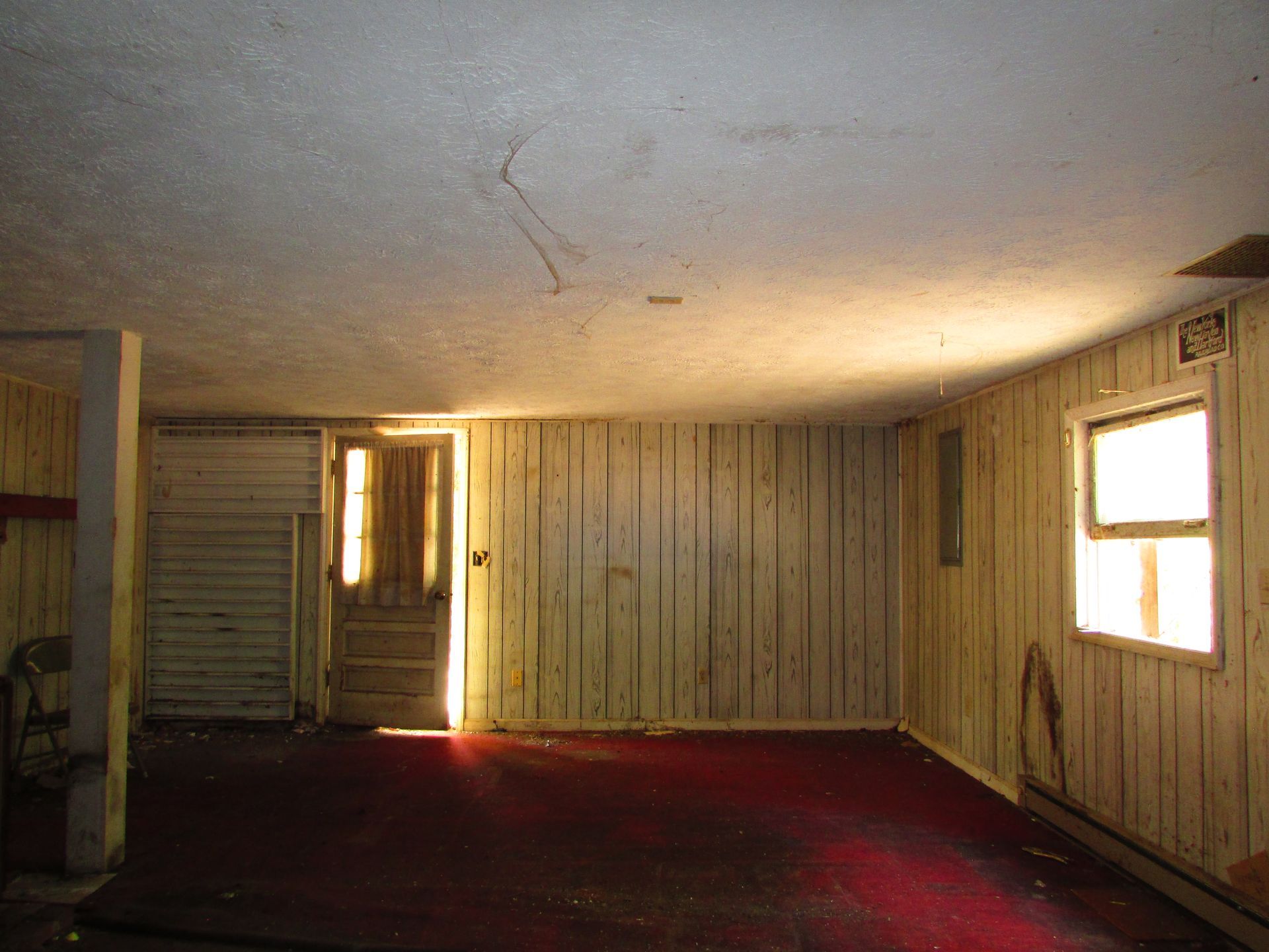 Empty, worn room with red carpet, wood-paneled walls, and two small windows.