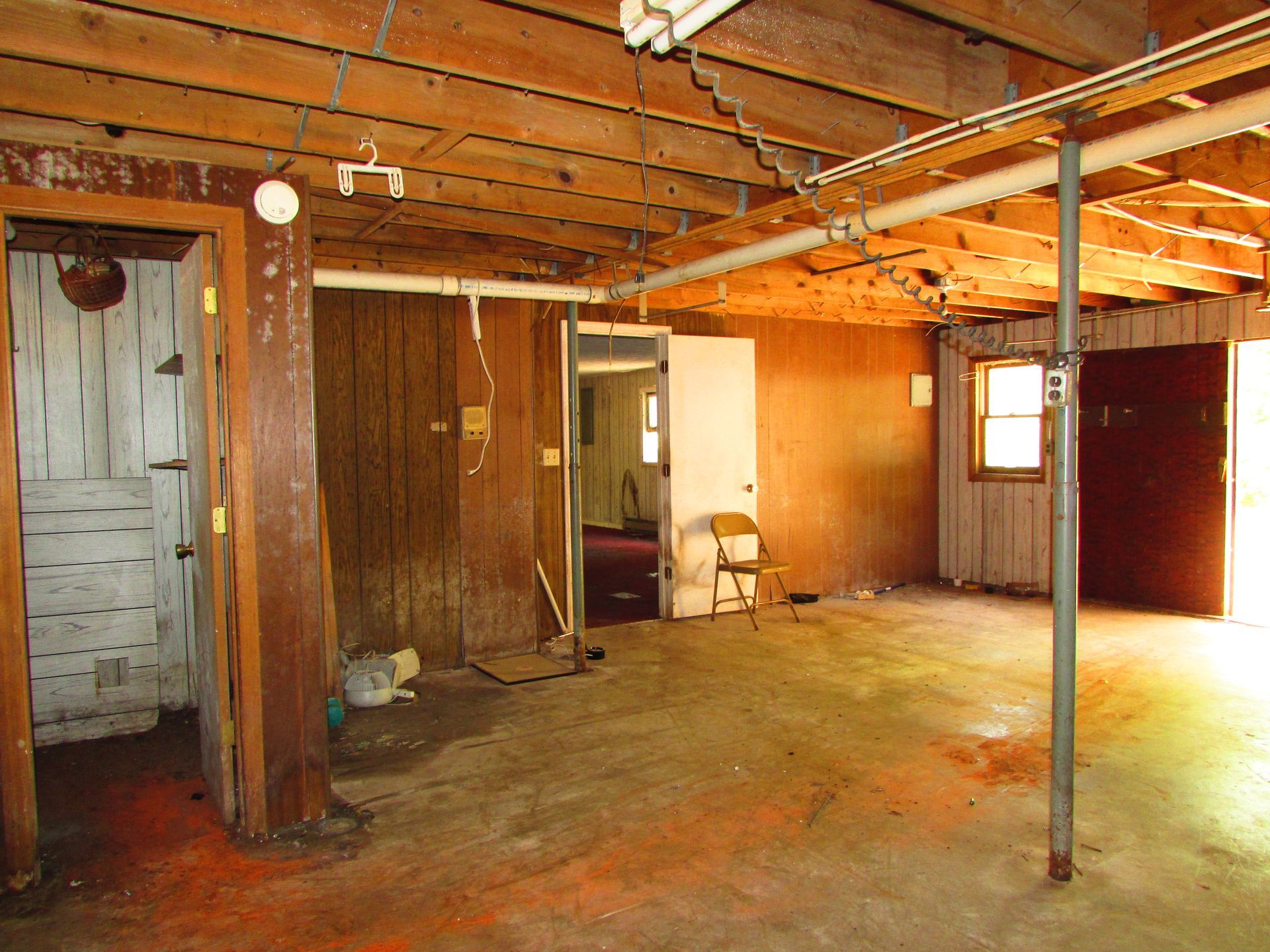 Empty unfinished basement with exposed beams, concrete floor, and a white door at left.