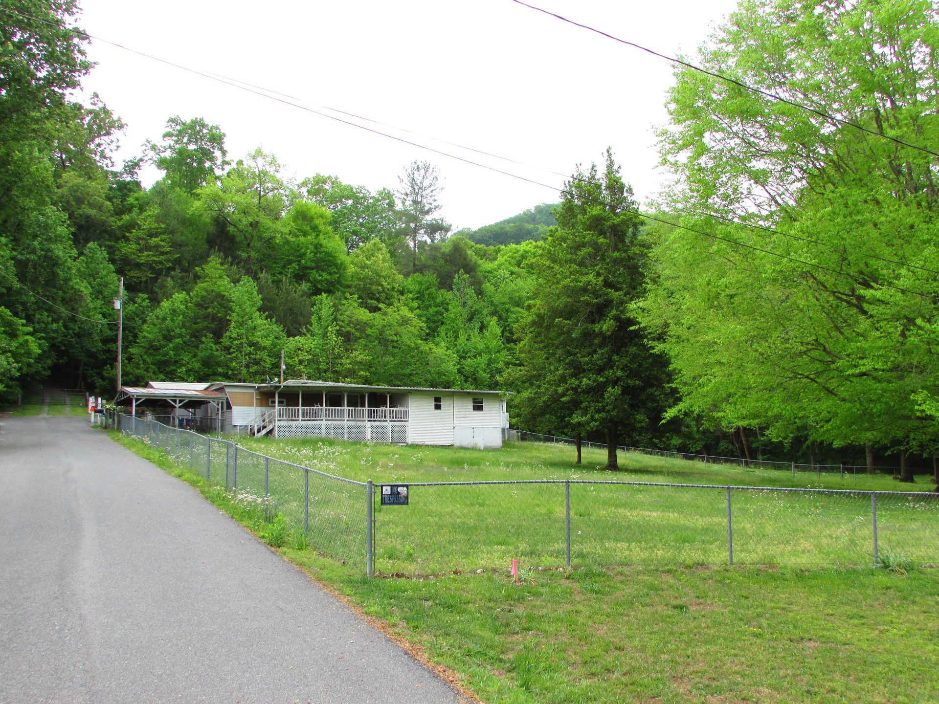 Rural roadside with a small white house, green trees, and a fenced grassy yard on a cloudy day