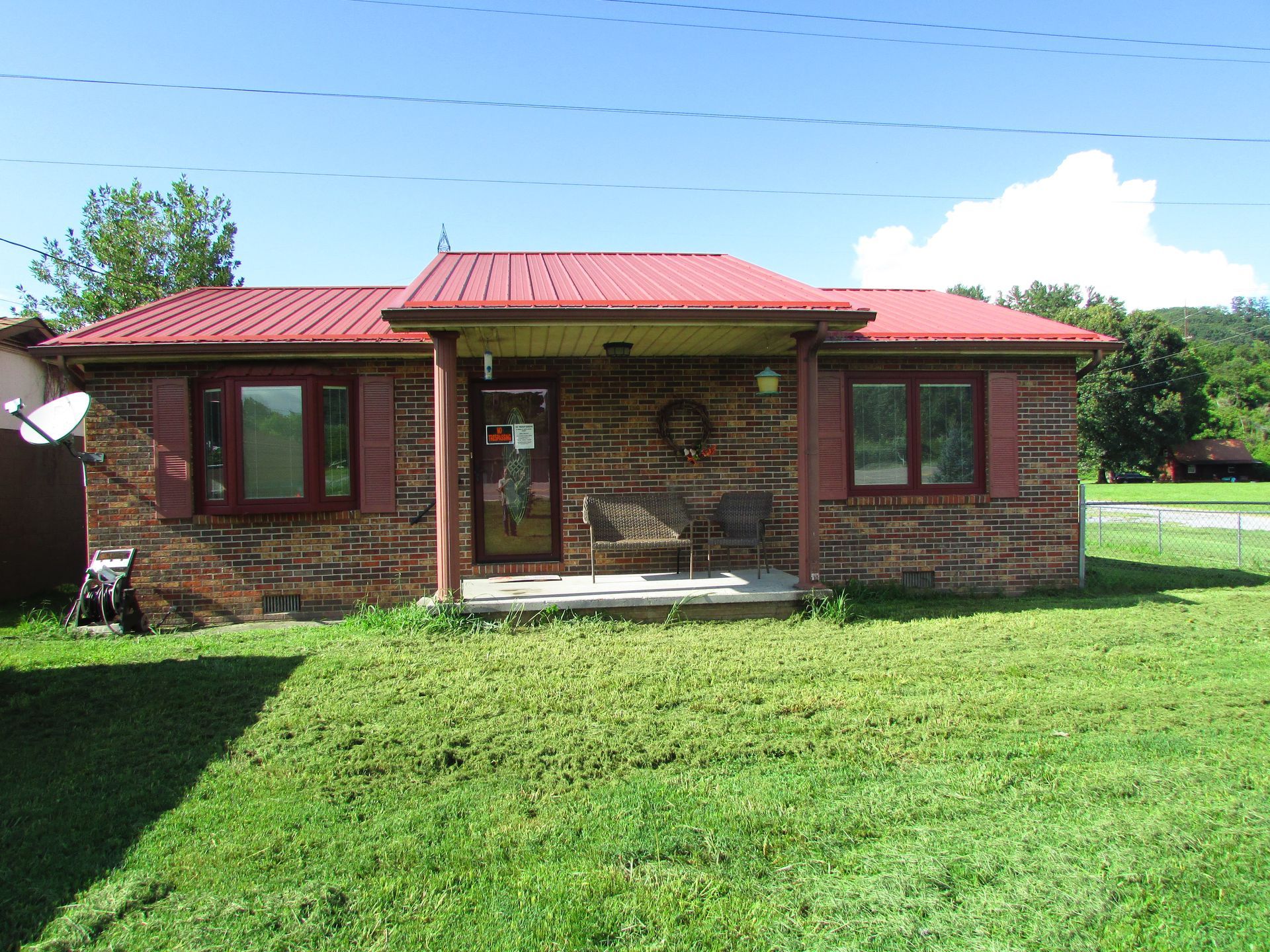 Small brick house with a red roof and front porch, set behind a green lawn under a blue sky.
