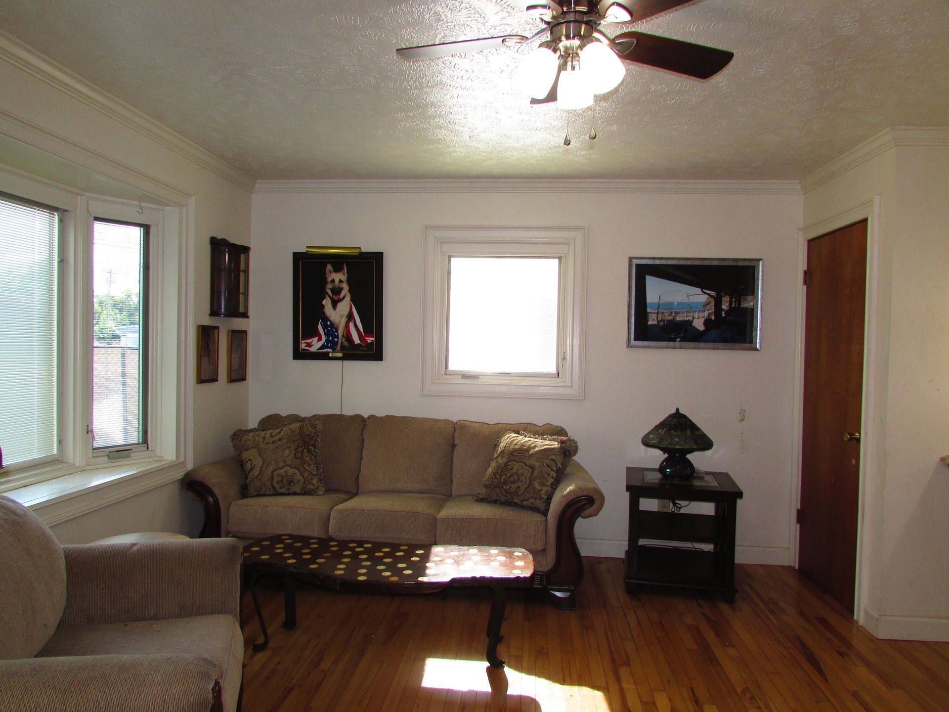 Living room with beige sofa, armchair, coffee table, wall art, and ceiling fan on hardwood floor.