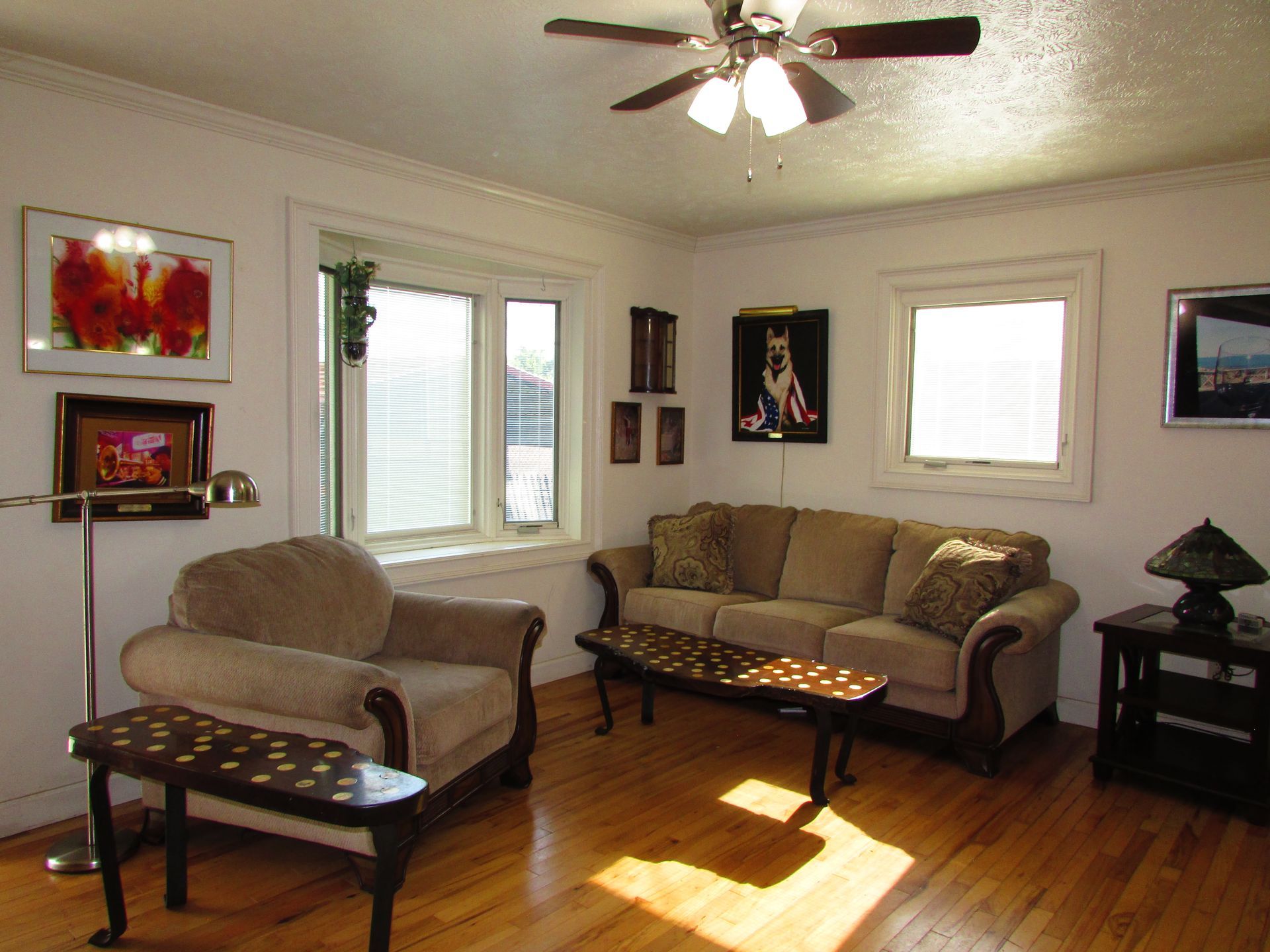 Bright living room with beige sofa and chair, hardwood floors, ceiling fan, windows, and wall art.