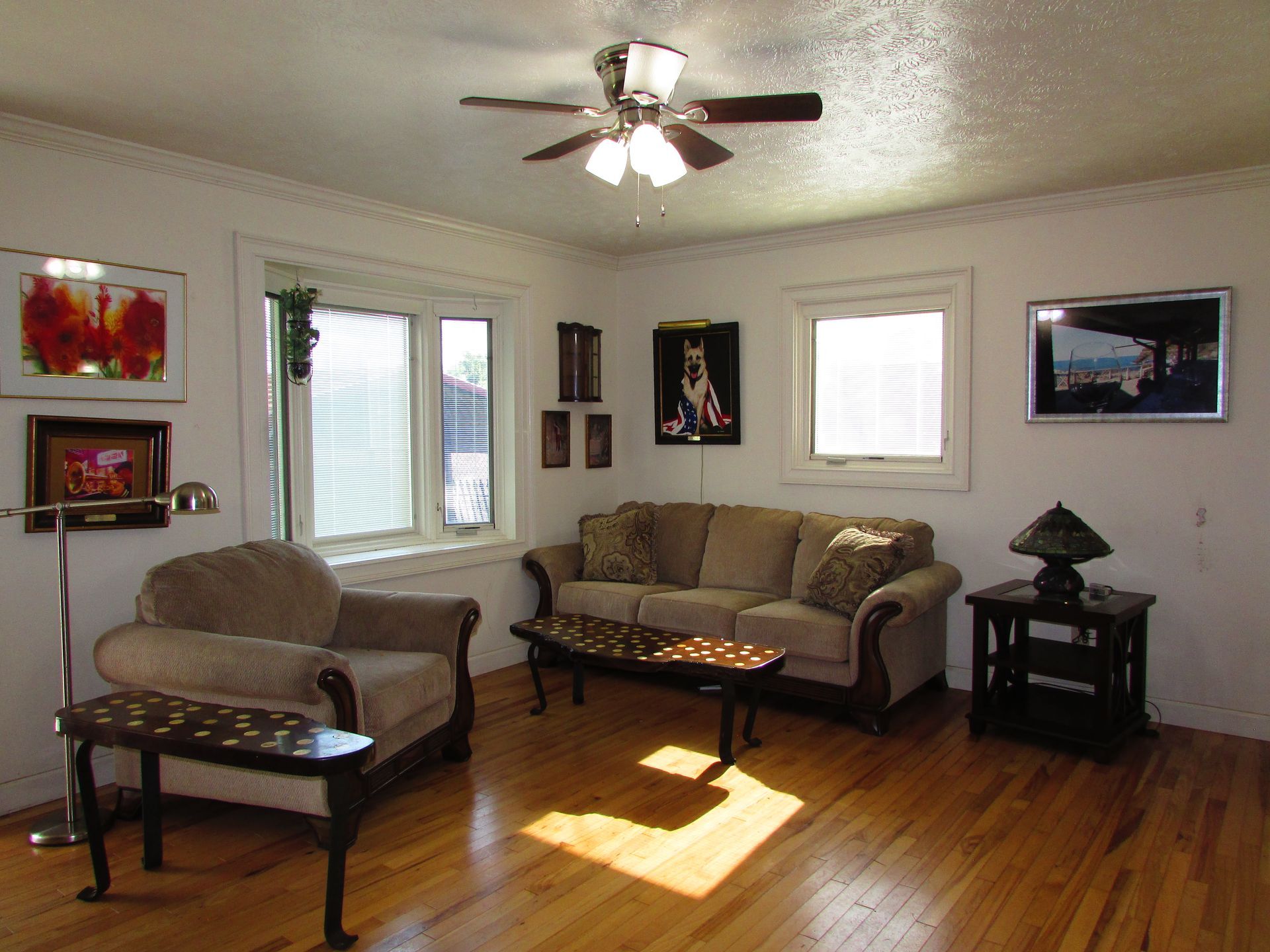 Living room with beige sofa and chair, coffee table, ceiling fan, framed art, and sunlight on hardwood floor
