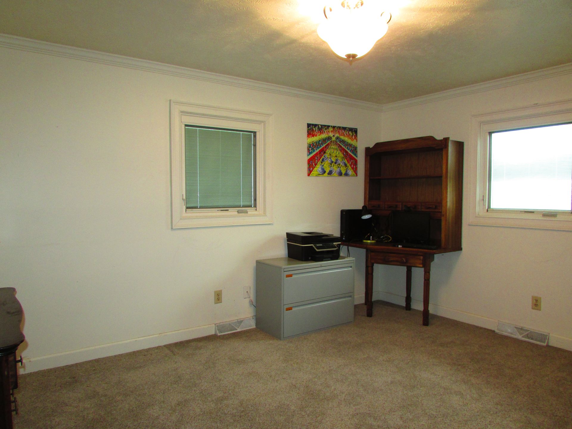 Empty beige bedroom with carpet, two windows, a dresser, and a small colorful wall hanging