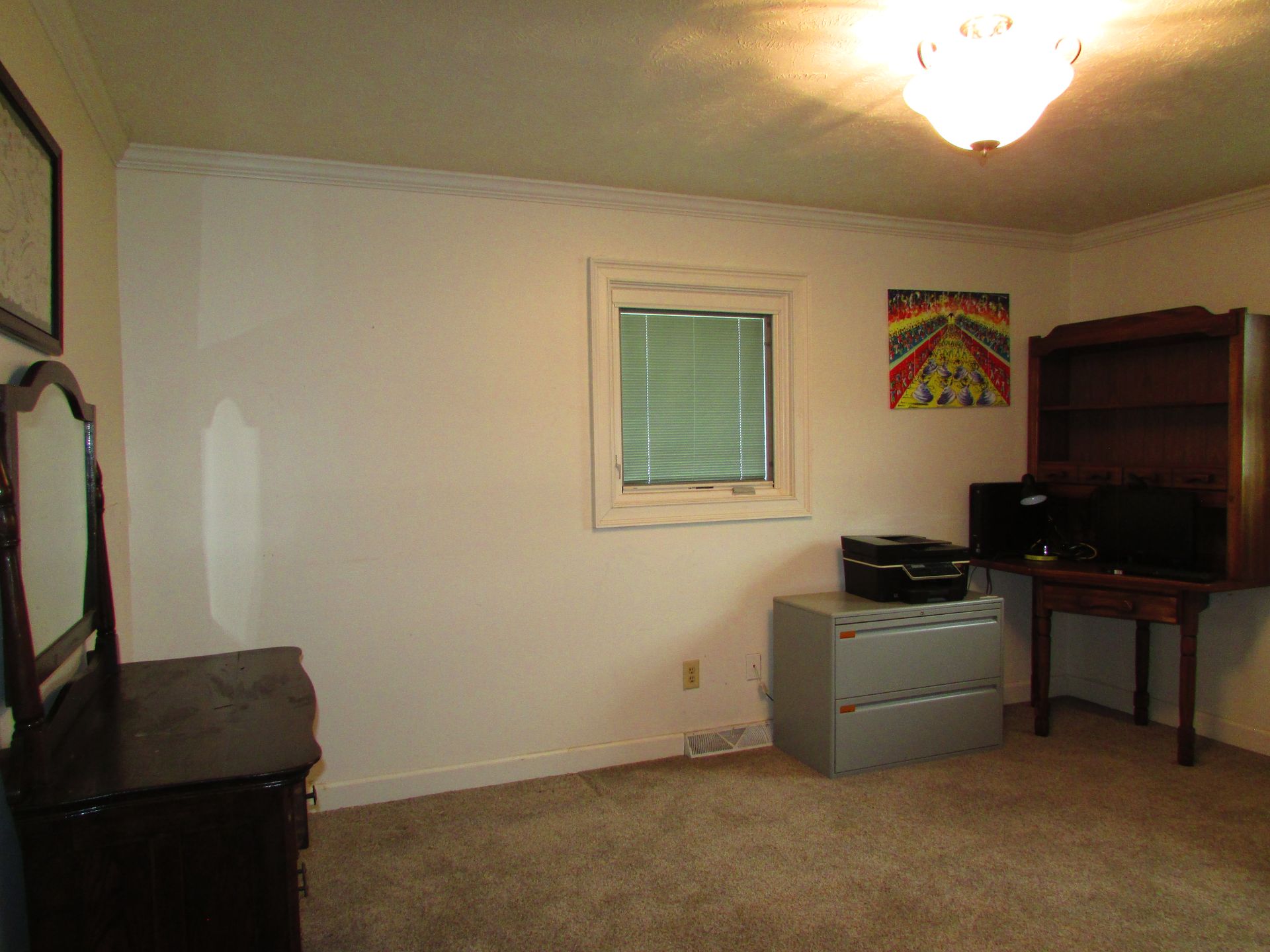 Empty bedroom with beige carpet, white walls, a window, and a desk, dresser, and bookshelf in the corner