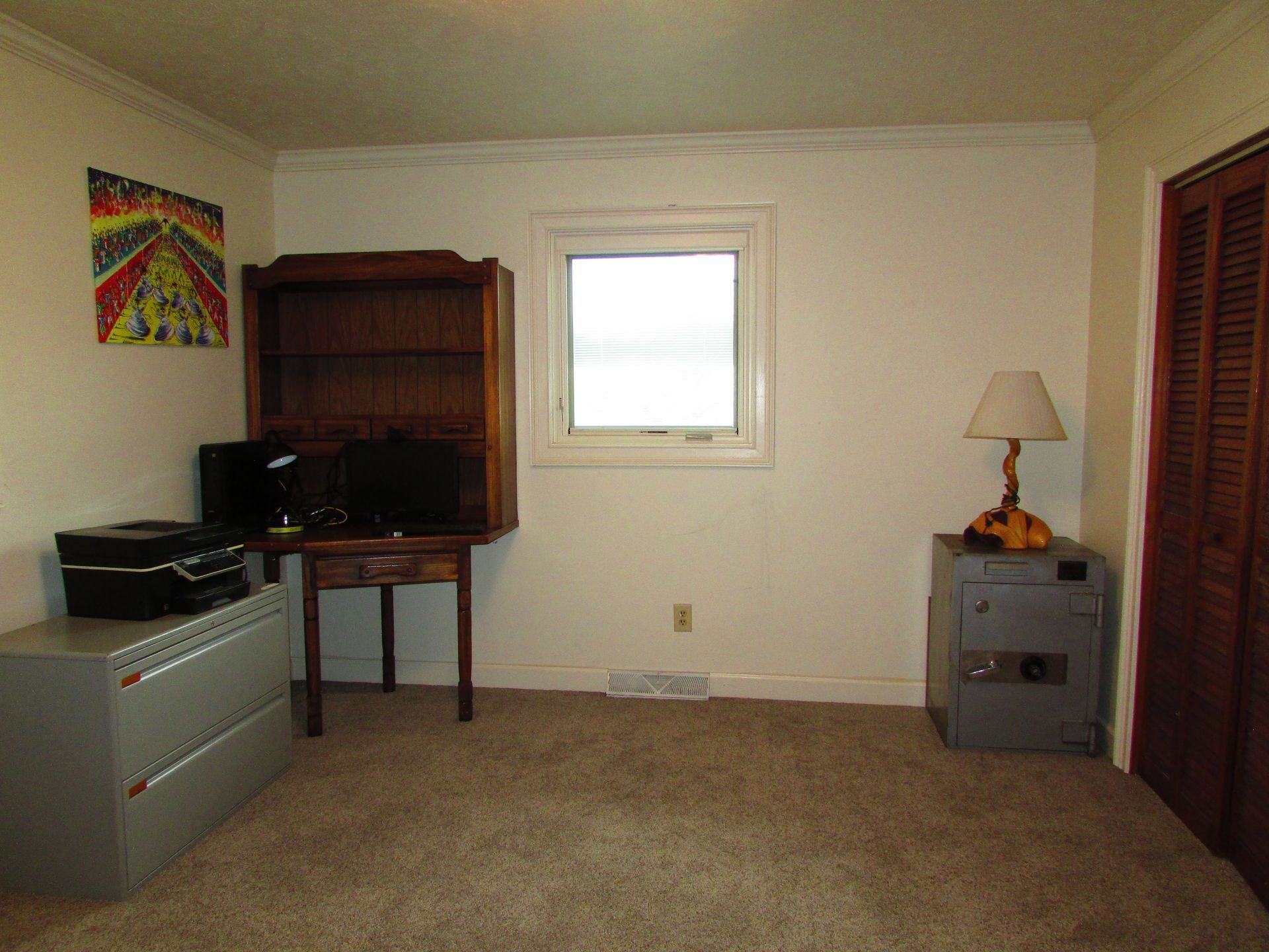 Empty beige room with carpet, window, desk, file cabinet, lamp, and red closet doors