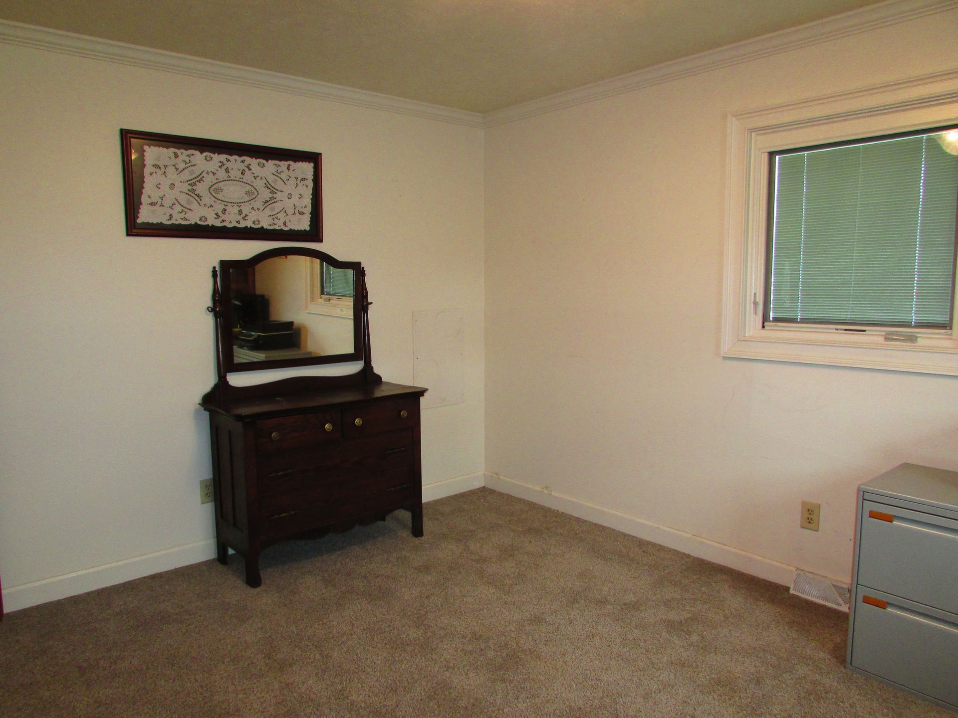 Empty beige bedroom with a dark dresser, wall mirror, window blinds, and carpeted floor.