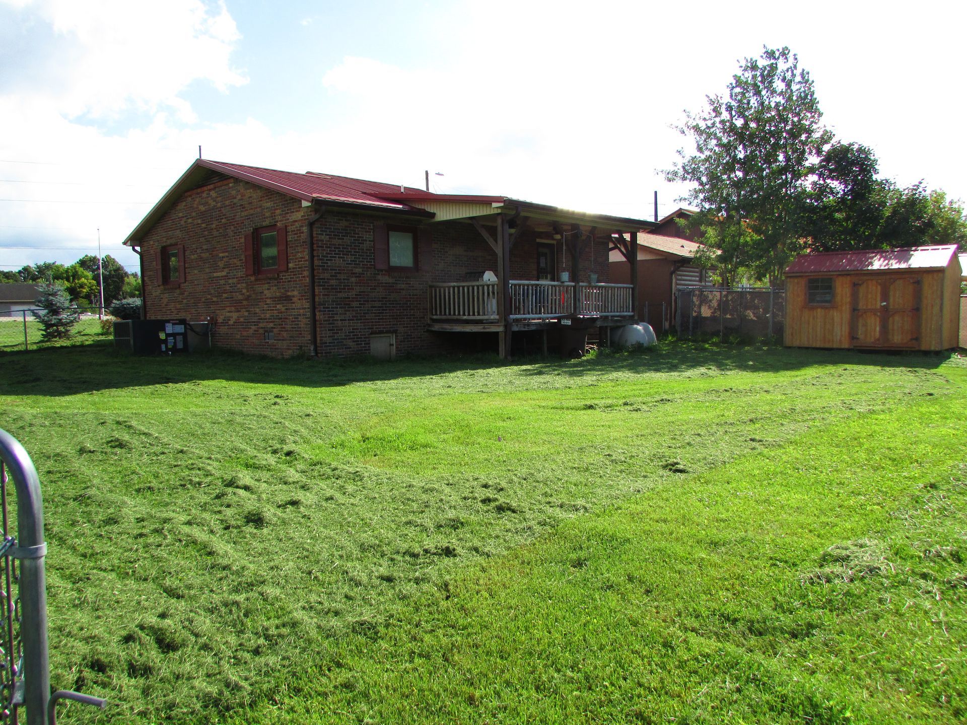 Brick house with red roof and porch in a sunny grassy yard, with a small shed and trees nearby