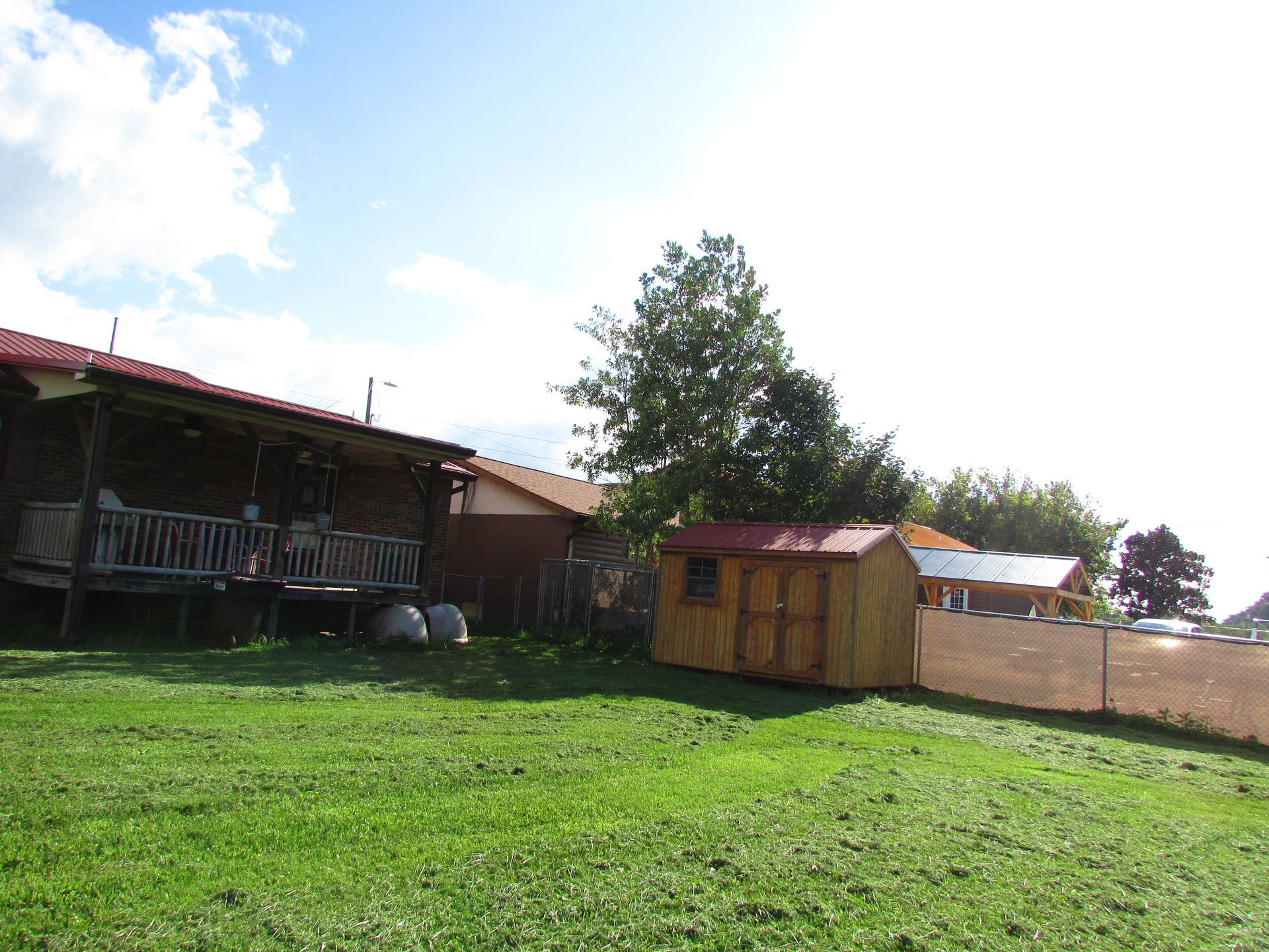 Rural yard with green grass, small wooden shed, house porch, and trees under a bright sky