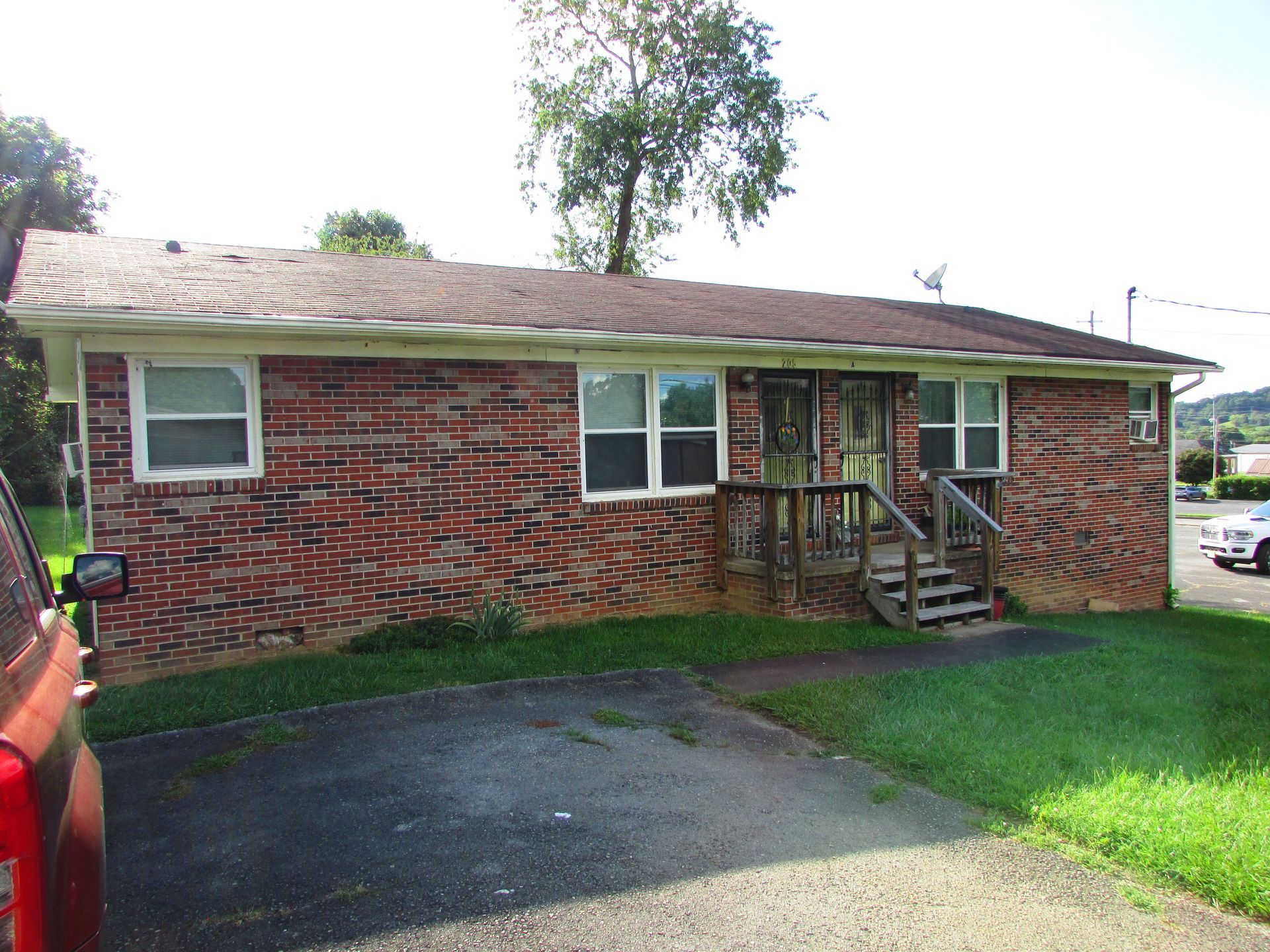 Single-story brick house with a front porch, driveway, and green lawn under bright sunlight