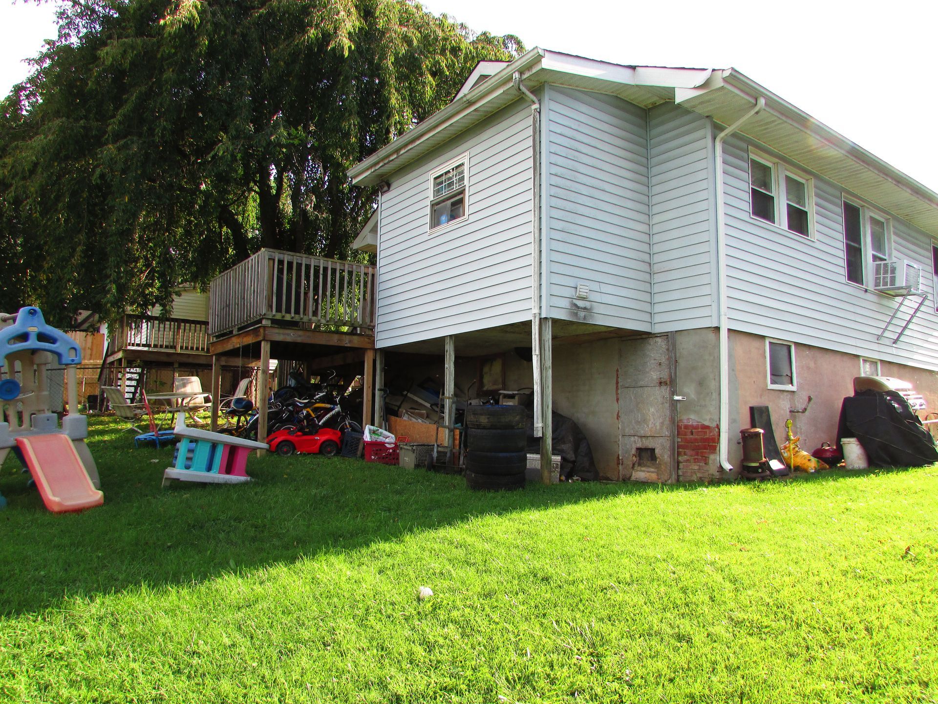 Backyard with a raised white house, green lawn, and scattered children’s toys near a tree