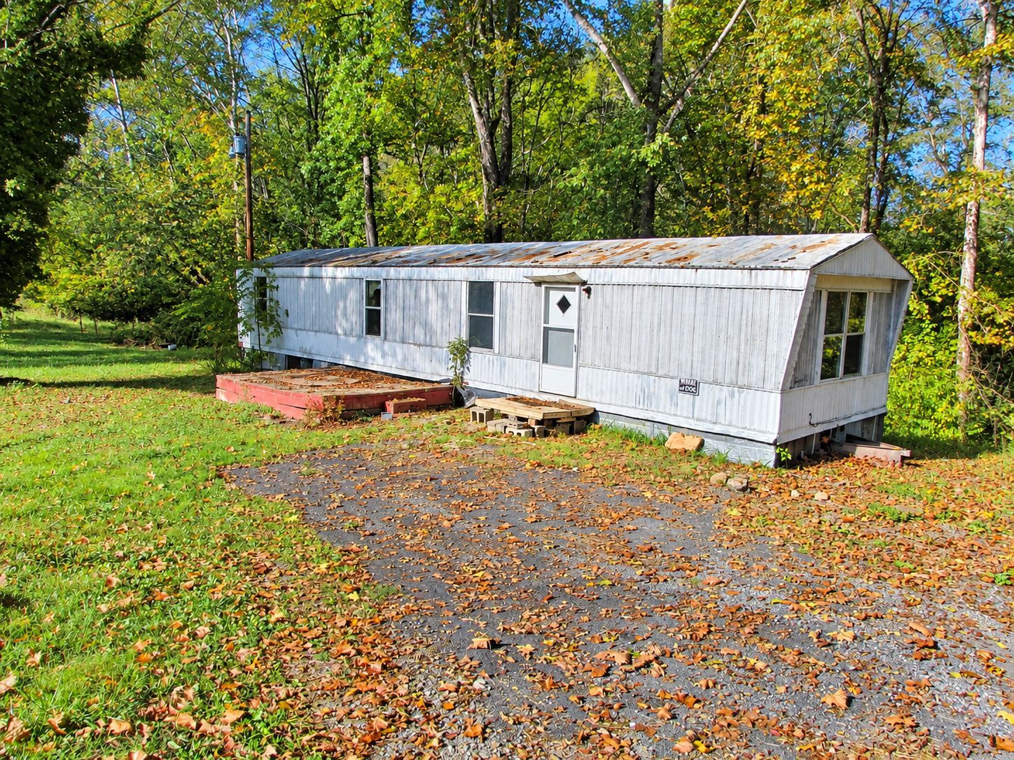 White mobile home with a porch on a gravel drive, surrounded by trees and autumn leaves.