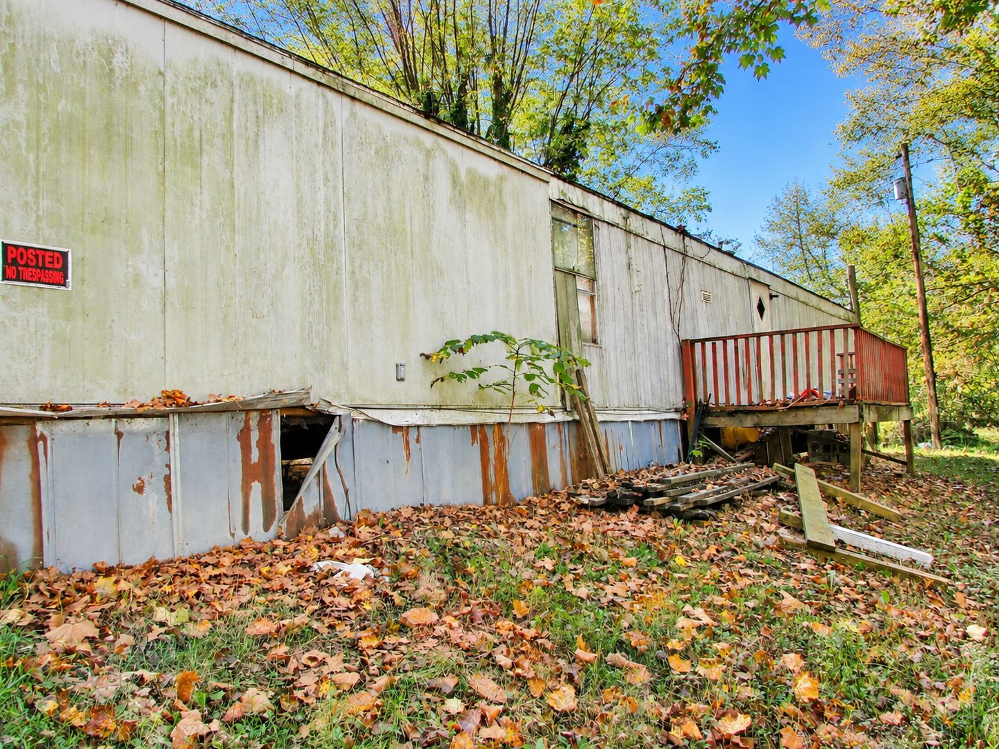 Abandoned mobile home with peeling white siding, rusted panels, and overgrown leaves in a wooded yard