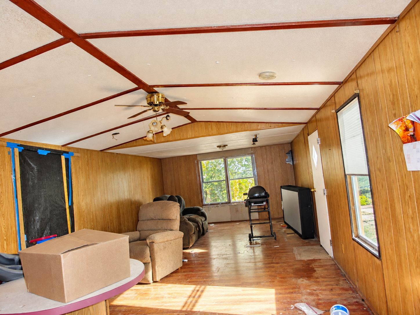 Small living room with wood-paneled walls, ceiling fan, chairs, boxes, and sunlight through windows.