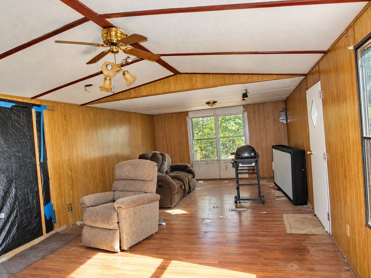 Empty mobile home interior with wood-paneled walls, recliner, ceiling fan, and a small stove by the window