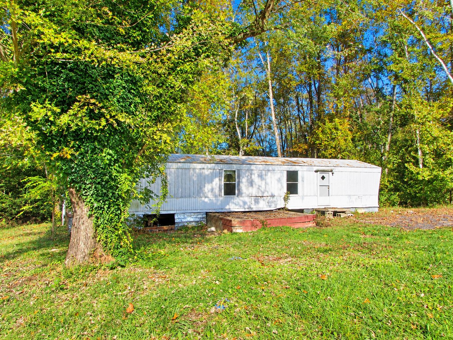 White mobile home beside a grassy hill, partially hidden by trees in a wooded yard